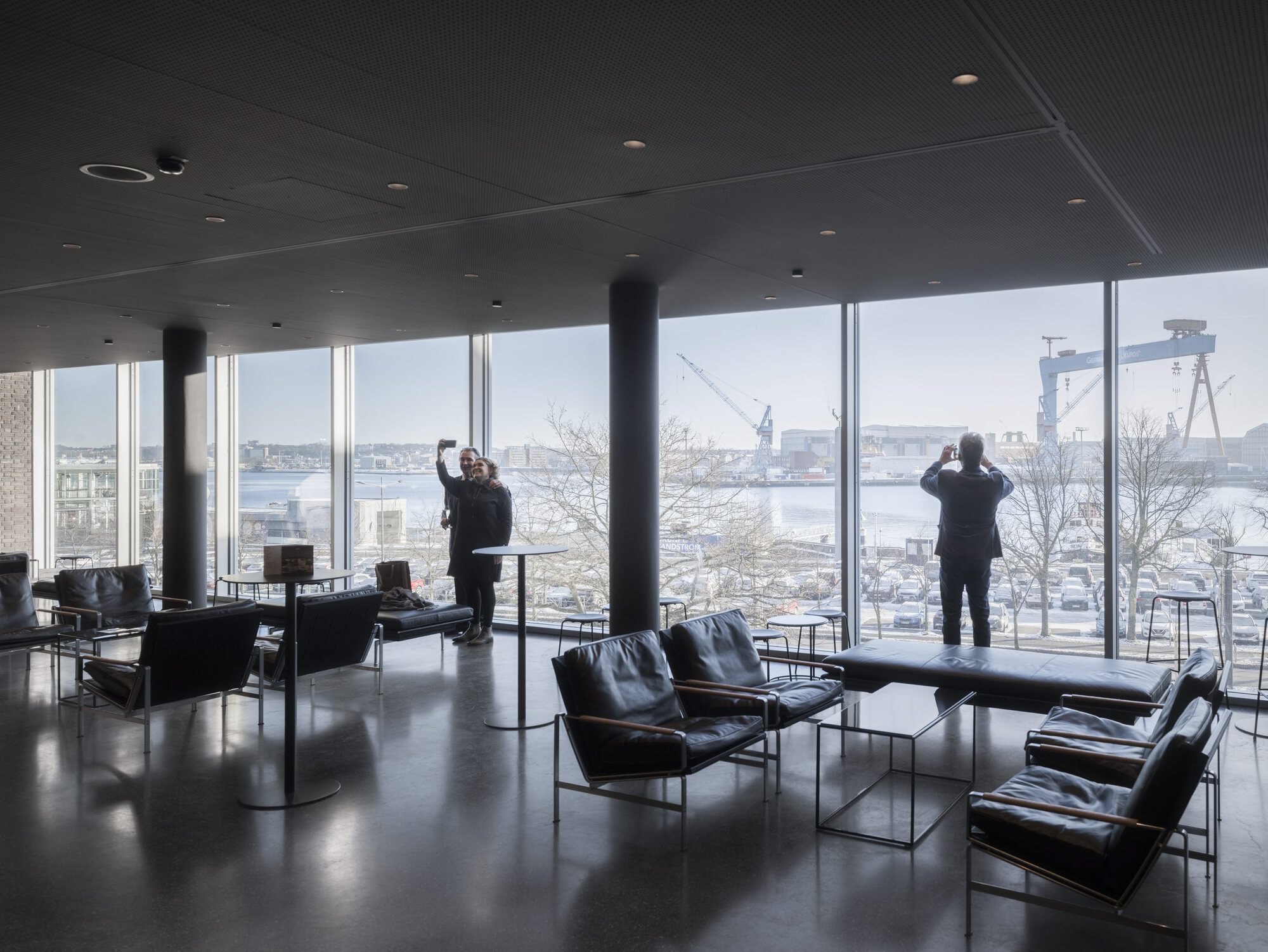 Interior lounge with black leather chairs and floor-to-ceiling windows overlooking industrial waterfront cranes