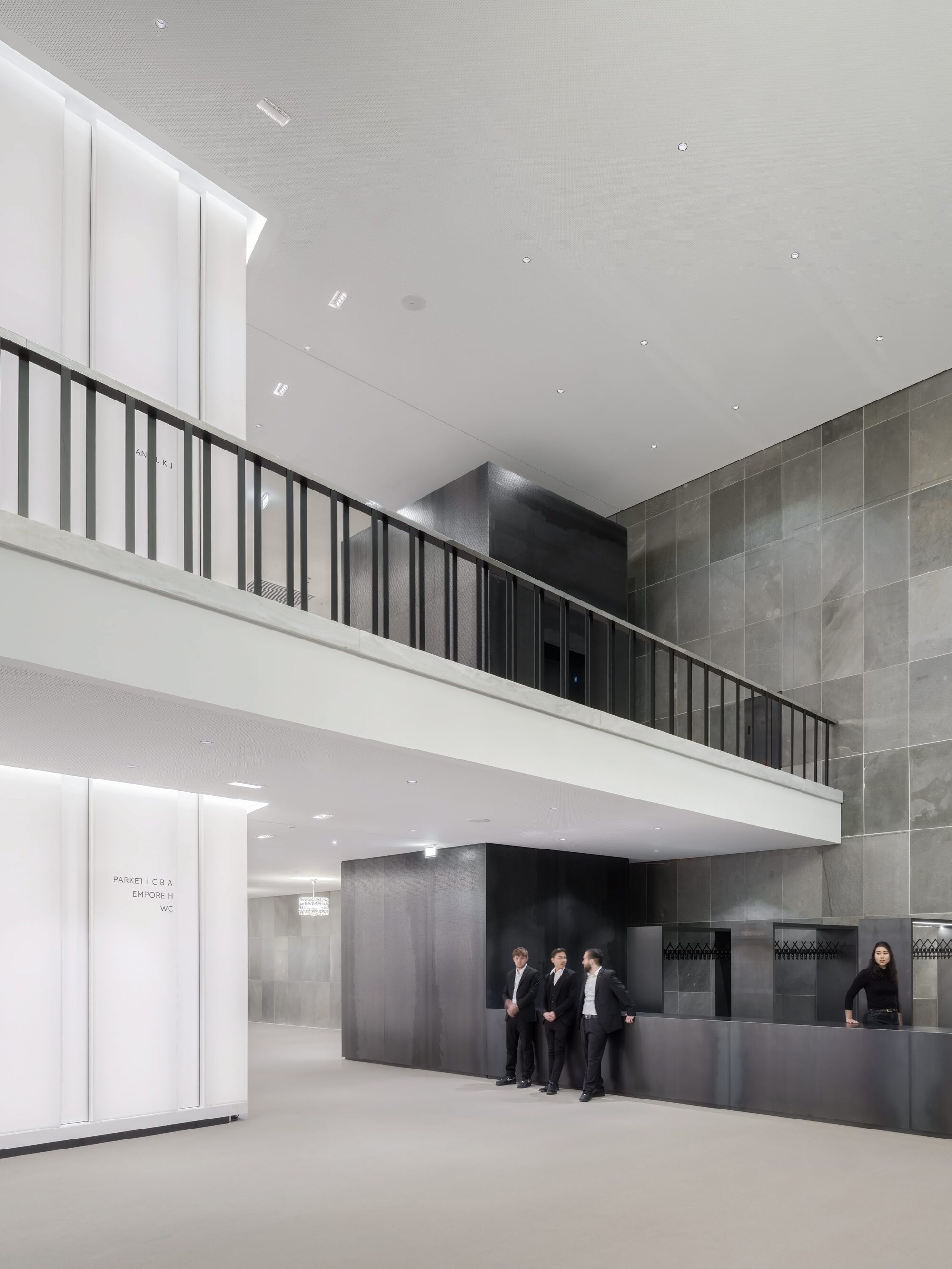 Double-height lobby space with black metal railing balcony and grey tiled wall beside reception desk
