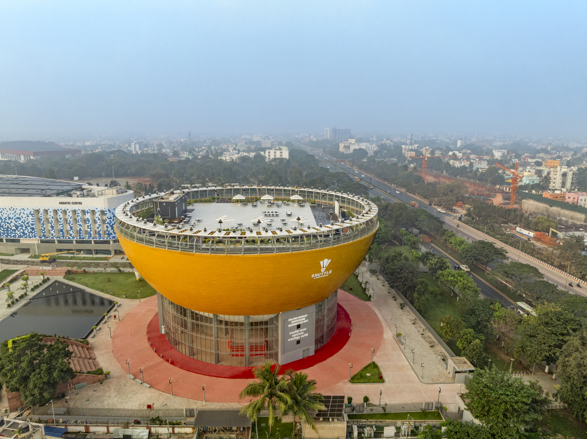 Drone perspective showing the circular rooftop plaza with glazed perimeter colonnade atop the yellow-clad bowl form