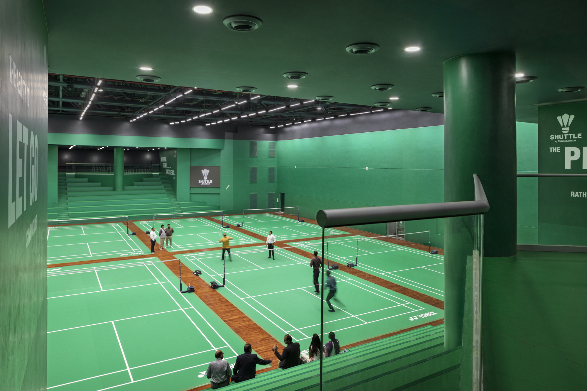 Interior view of green-walled badminton courts with players and spectators beneath recessed ceiling lighting