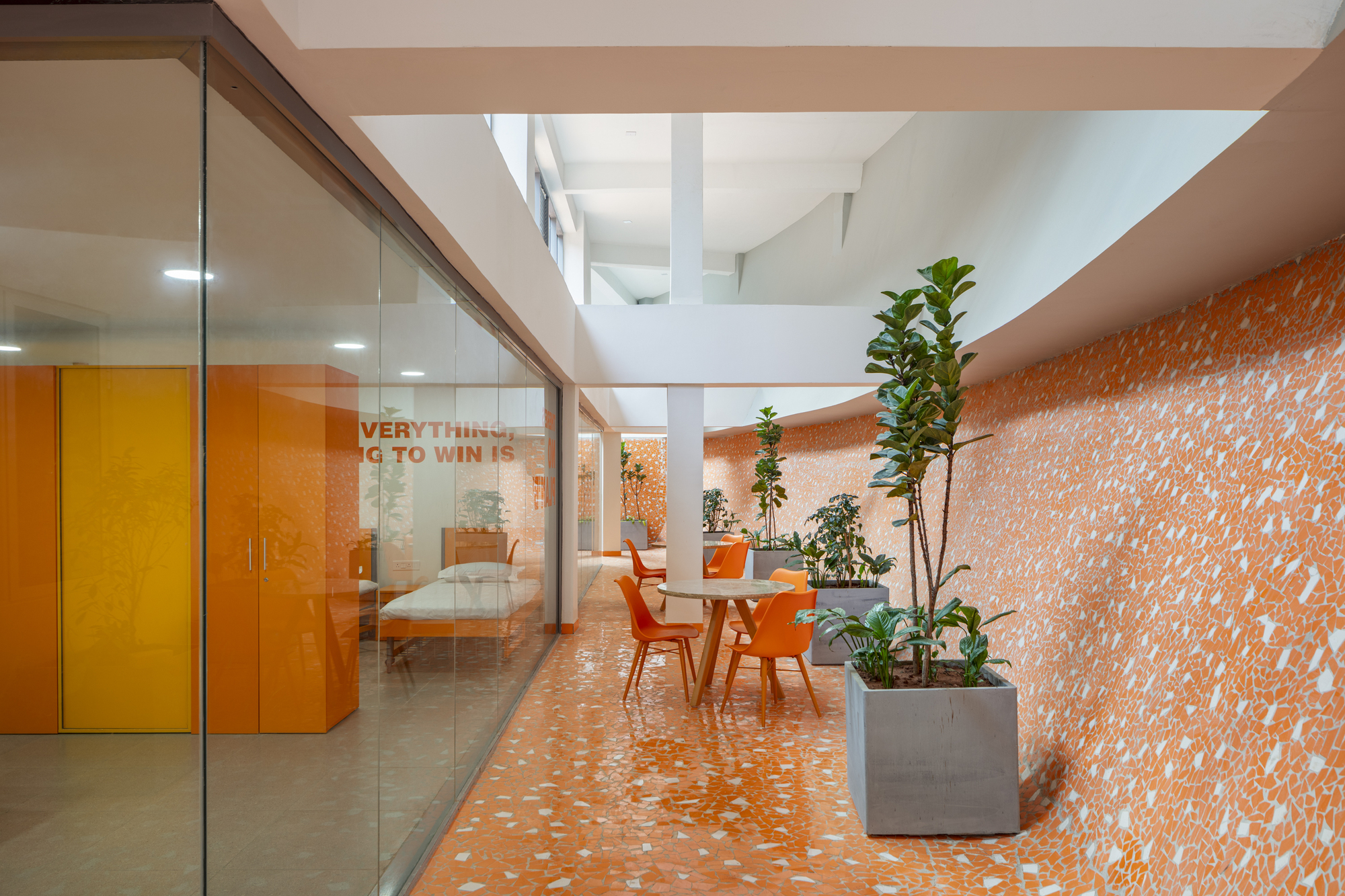 Interior corridor with orange terrazzo flooring, glazed office partitions, potted plants and orange seating under skylights