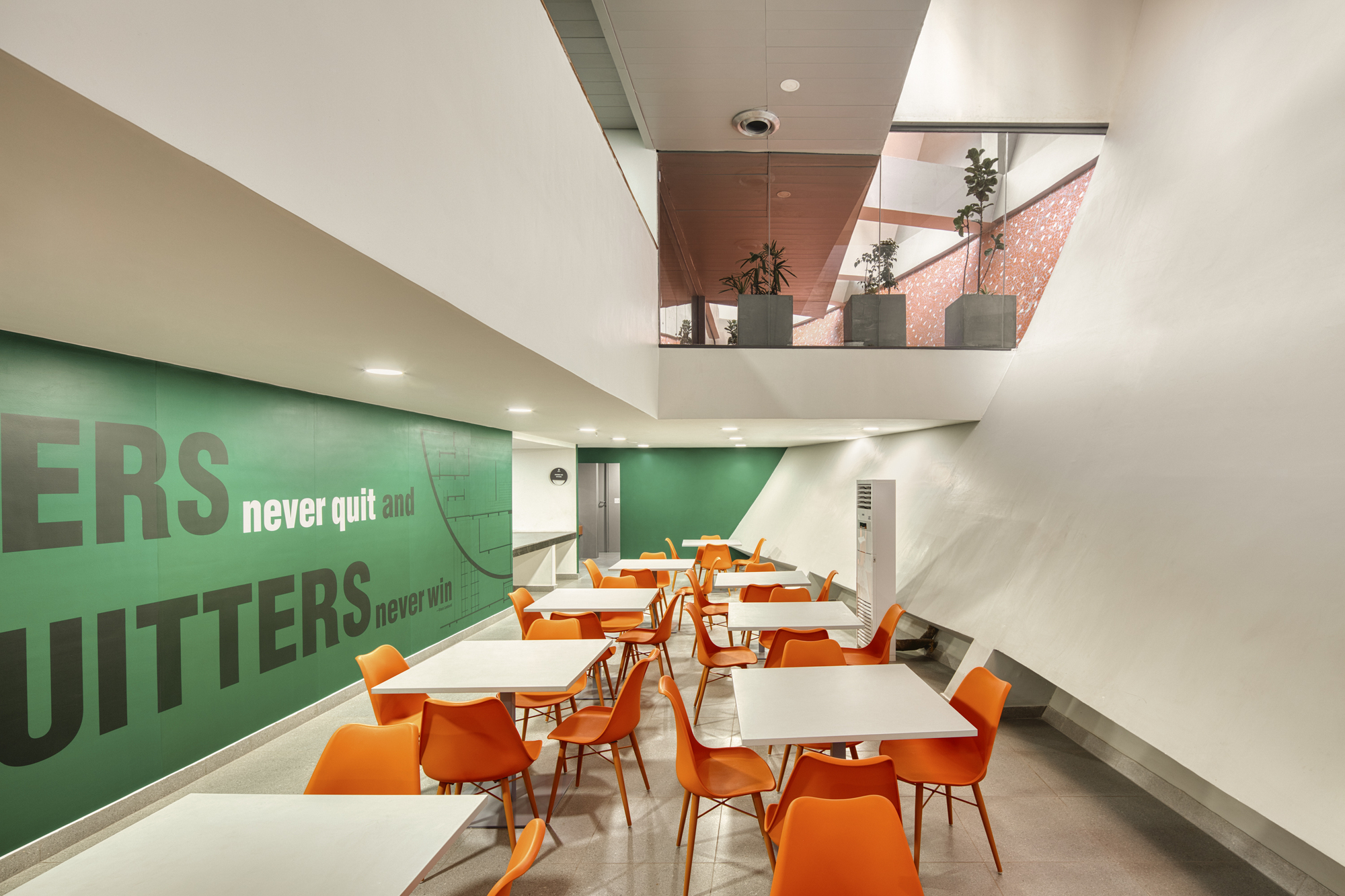 Interior dining area with orange chairs and green wall graphics beneath a skylit mezzanine with planters