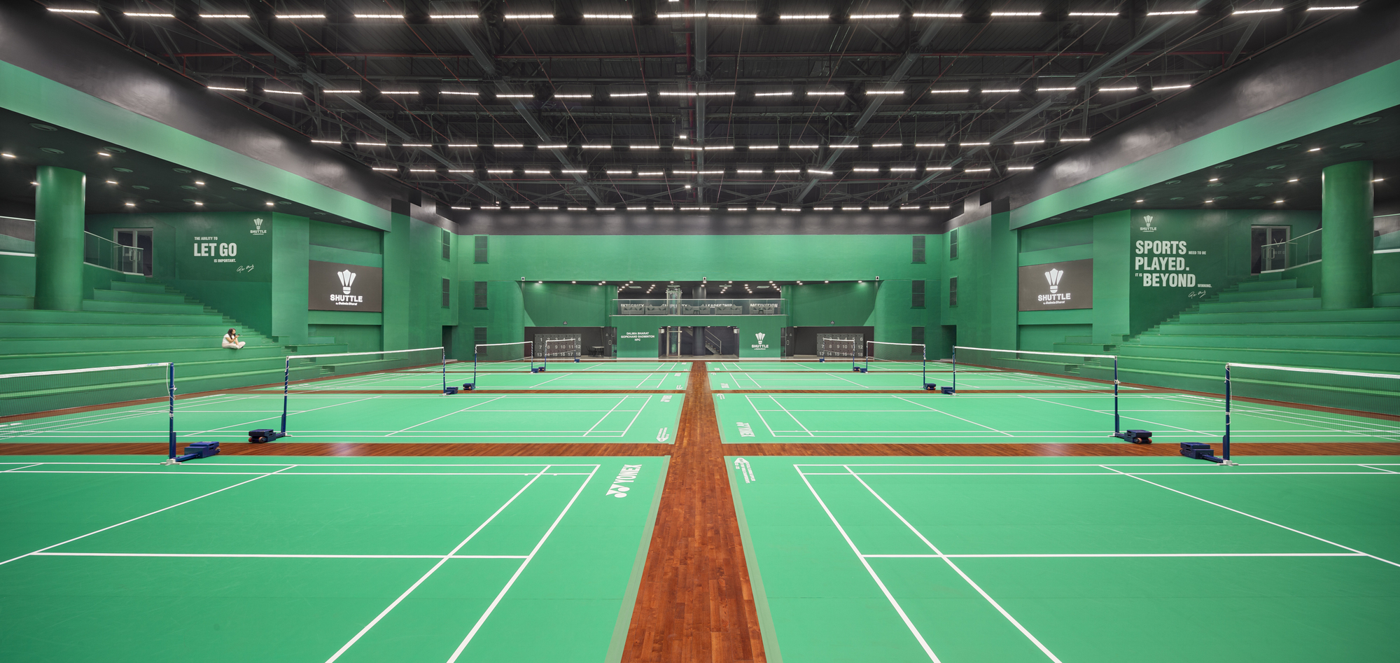 Badminton courts with green playing surface and mint walls under a black ceiling grid with linear lighting
