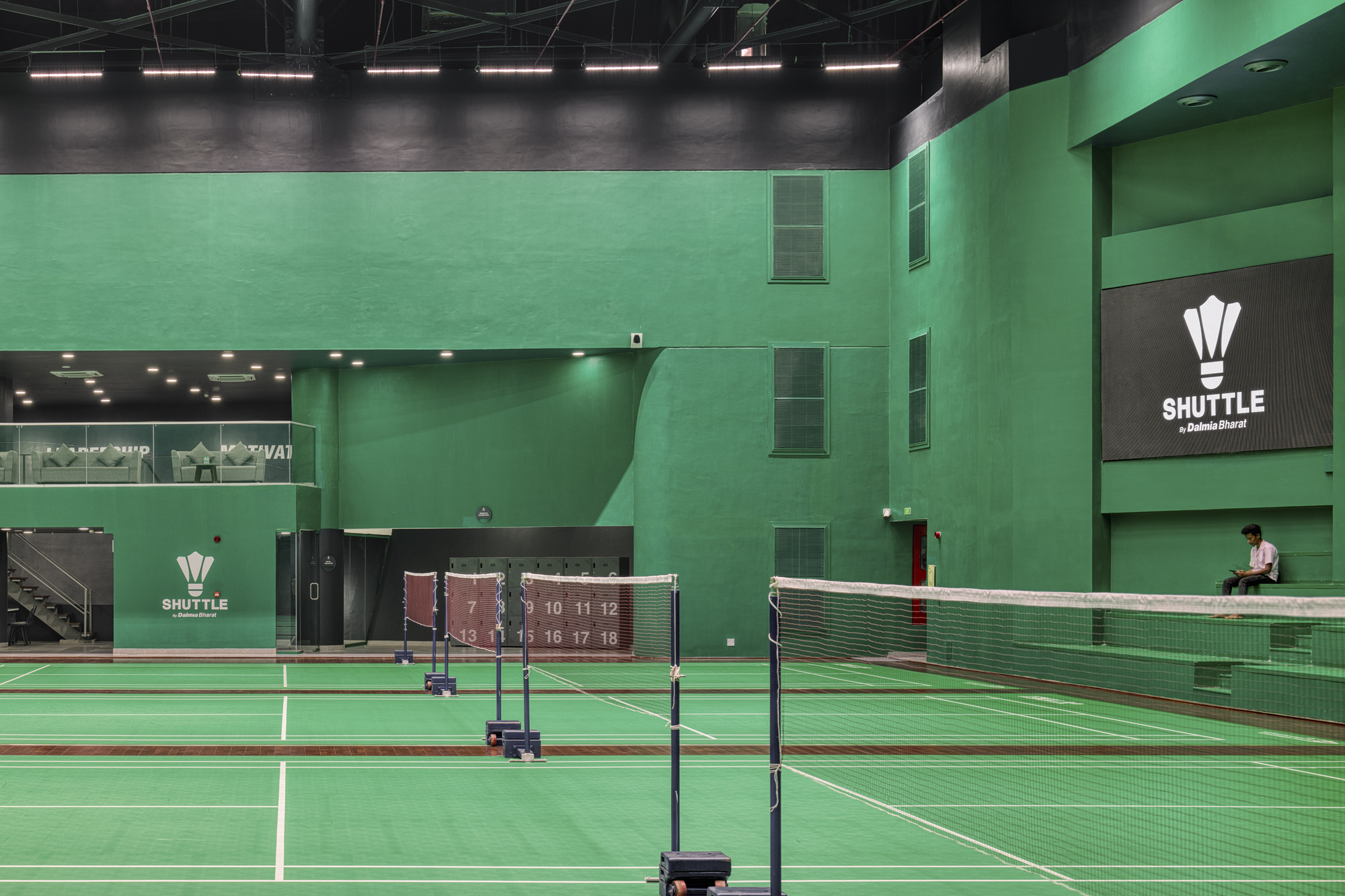Multi-level sports hall interior with green walls, badminton nets, and a lone figure on the upper level