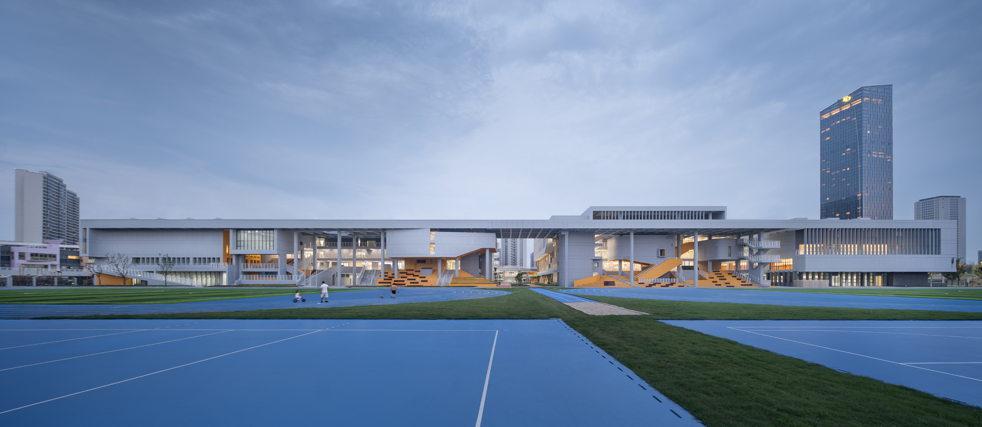 Horizontal facade with illuminated glazing behind a blue running track and grass field at dusk