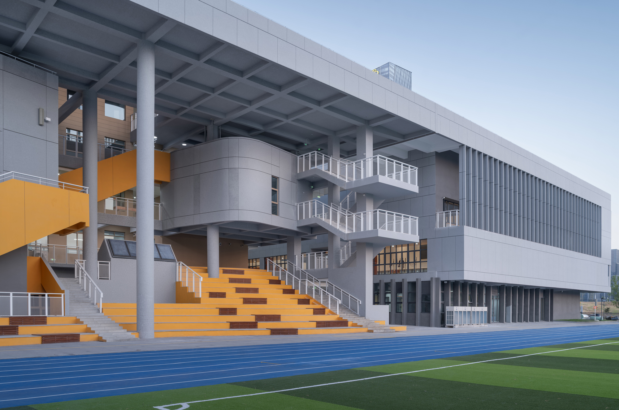Orange tiered seating stairs beneath deep overhang beside blue athletic track and green sports field