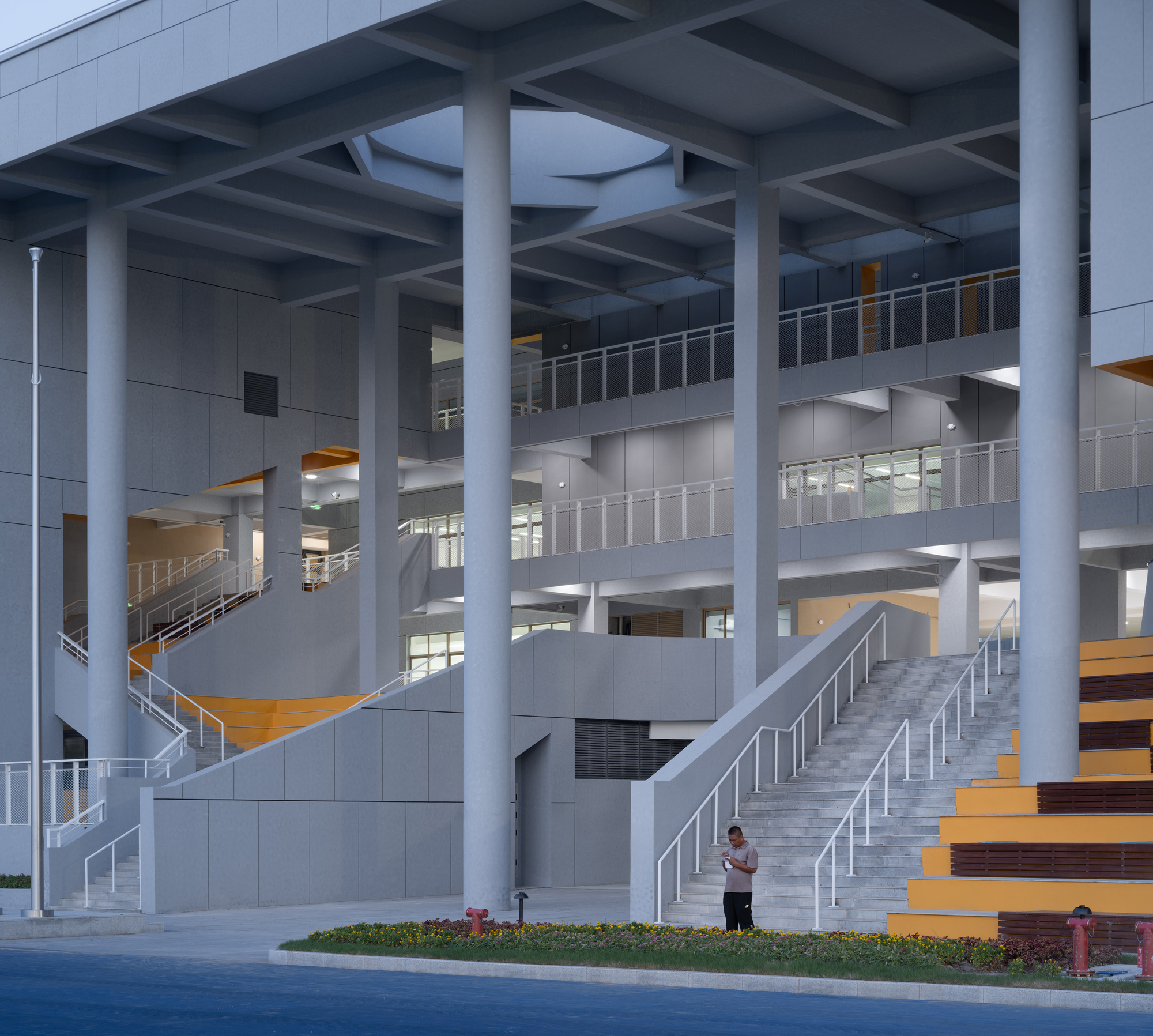 Covered ground level atrium with grey concrete columns, timber-lined staircases, and a pedestrian walking beneath