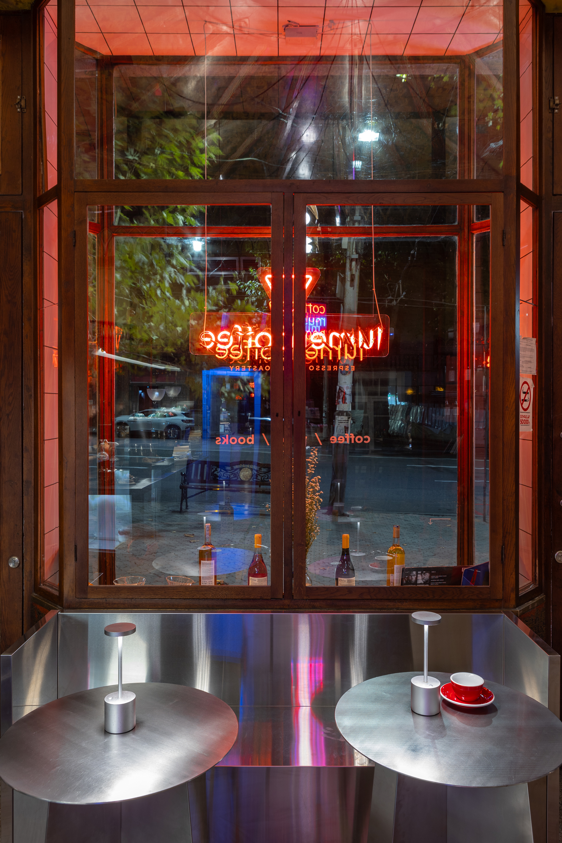 Glass storefront with neon signage reflected in the window above two circular steel tables at night