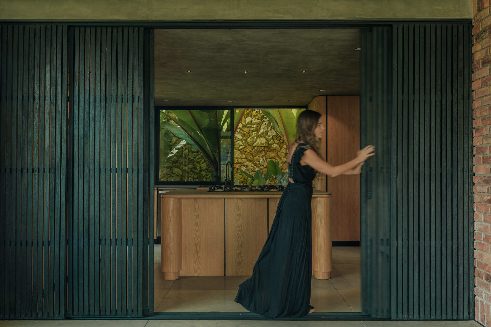 Kitchen view through folding dark green slatted panels as a woman in evening attire opens a timber door