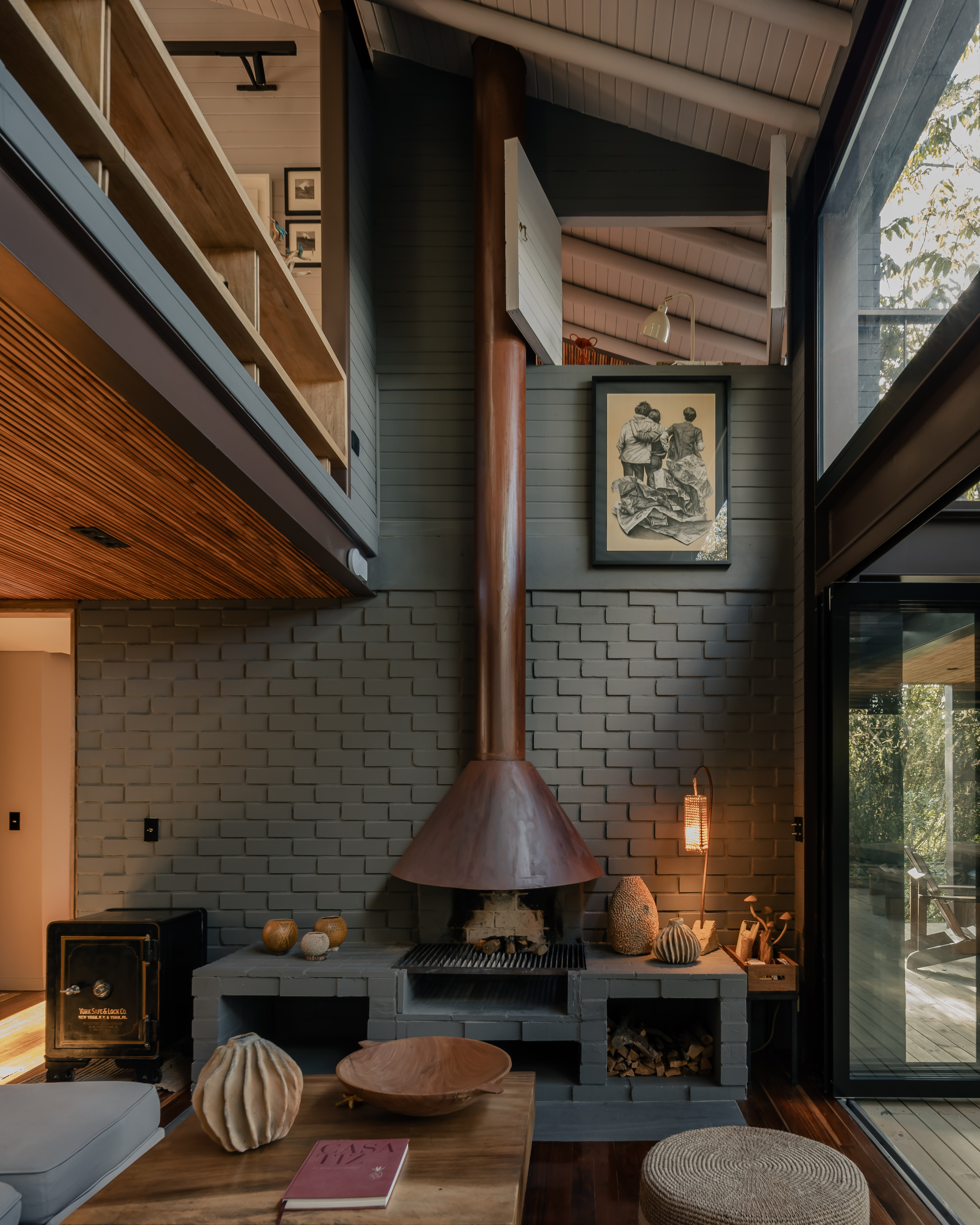 Freestanding copper fireplace hood above concrete plinth with timber ceiling and framed artworks at dusk