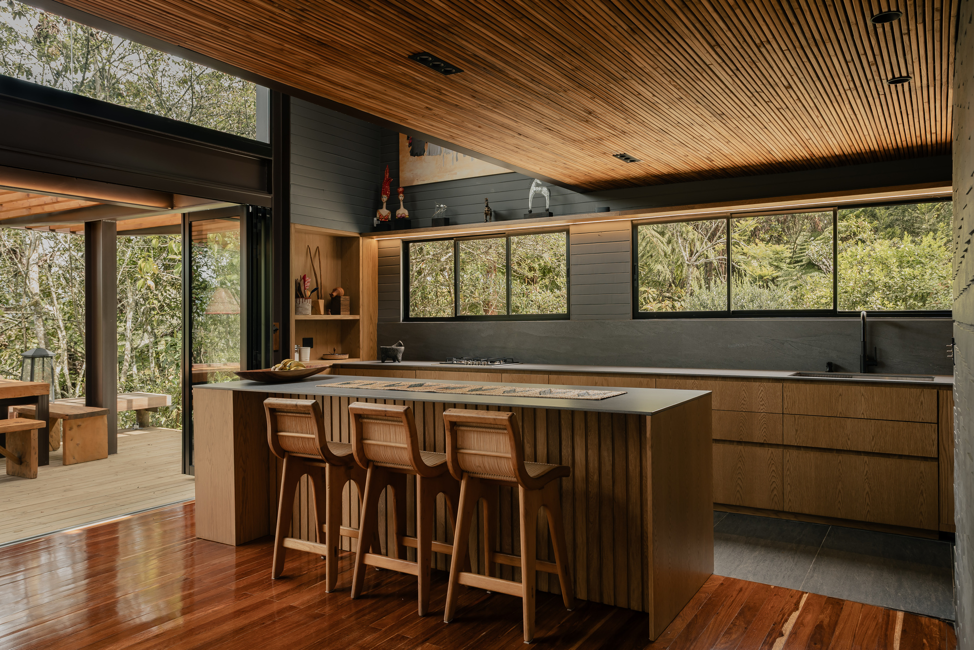 Kitchen island with timber cabinetry and slatted ceiling overlooking forested hillside through clerestory glazing