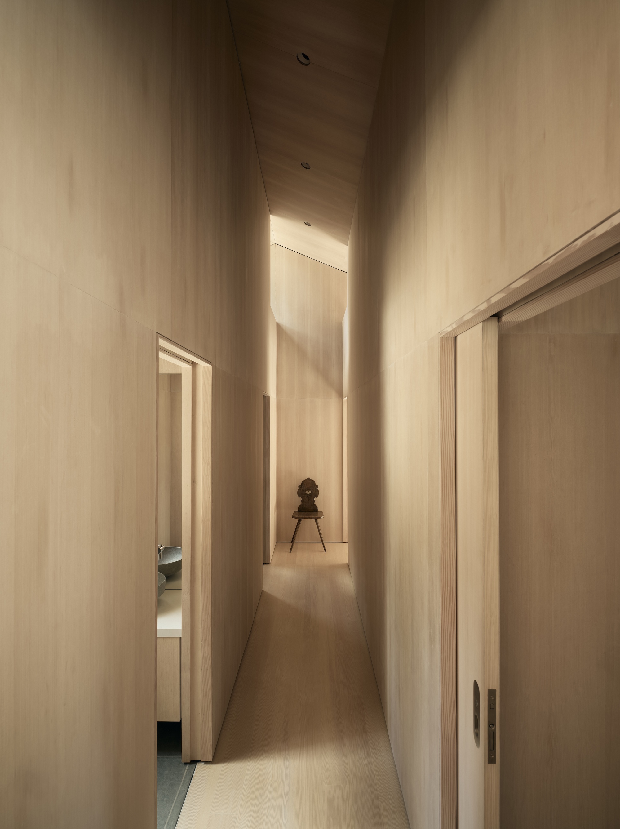 Narrow corridor with light wood walls and ceiling framing a small chair at the end