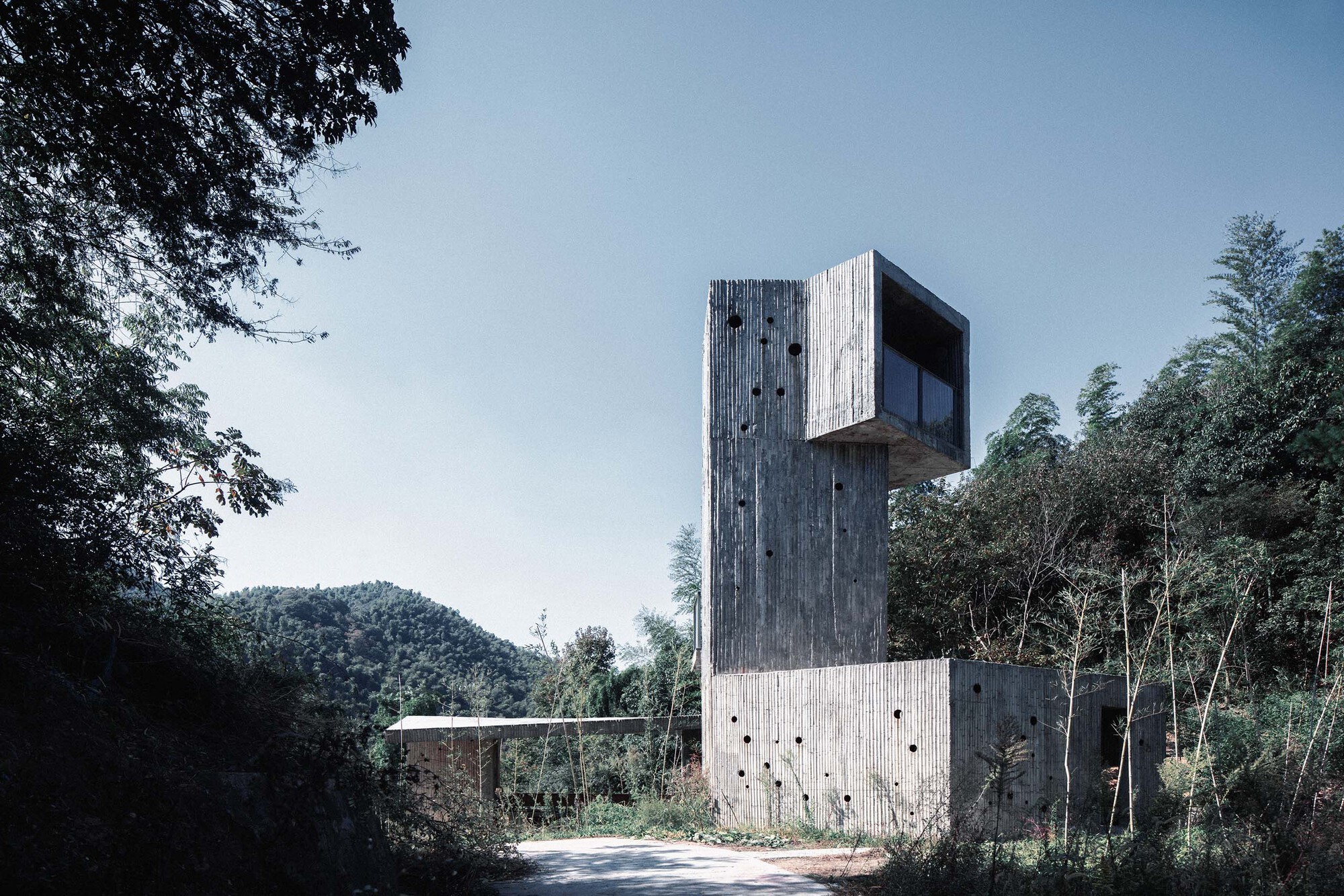 Vertical concrete tower with board-formed walls rising above a wooded hillside road