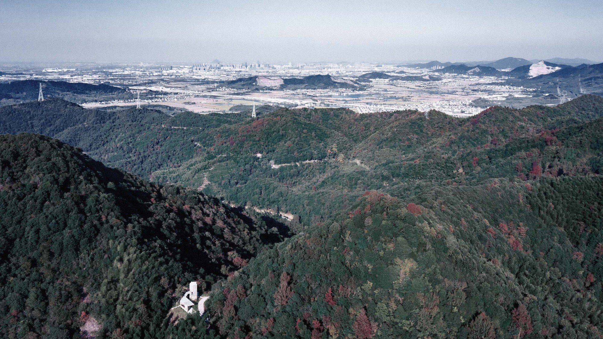 Drone view of the tower on a forested ridge with distant city sprawl under hazy sky