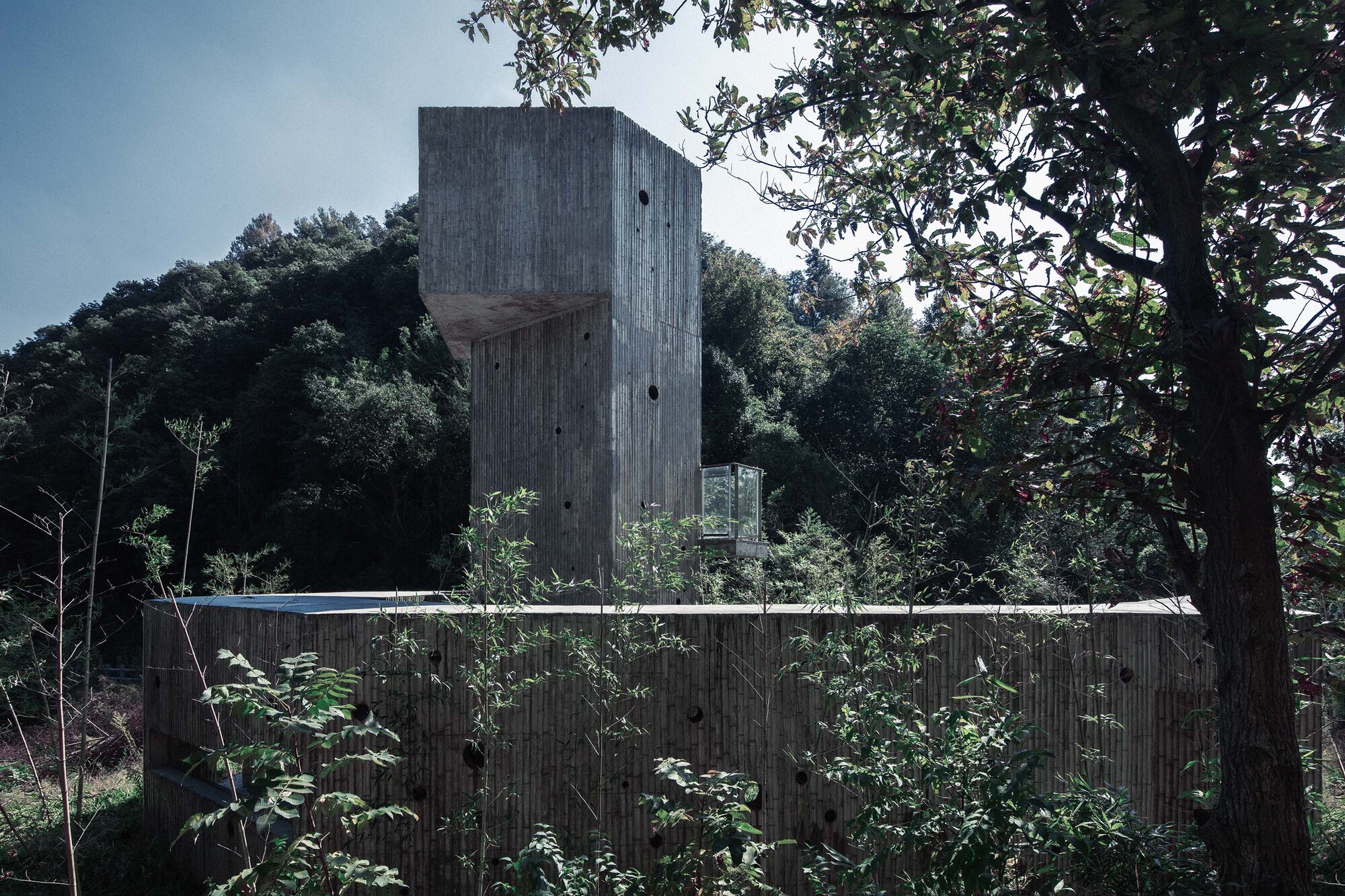 Vertical concrete tower behind a horizontal timber-clad fence partially obscured by vegetation