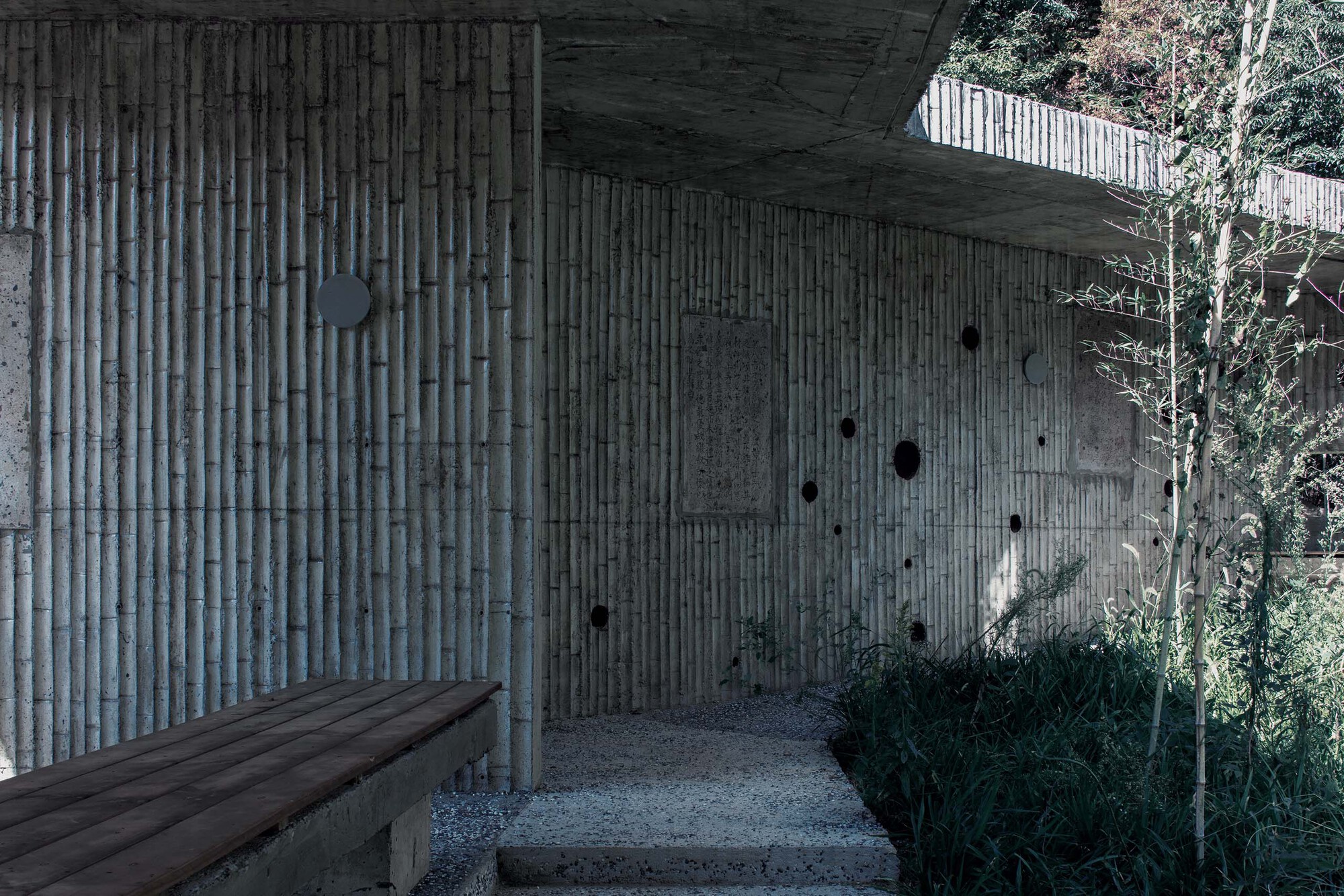 Covered entry courtyard with vertical timber formwork walls and gravel floor surrounded by planted vegetation