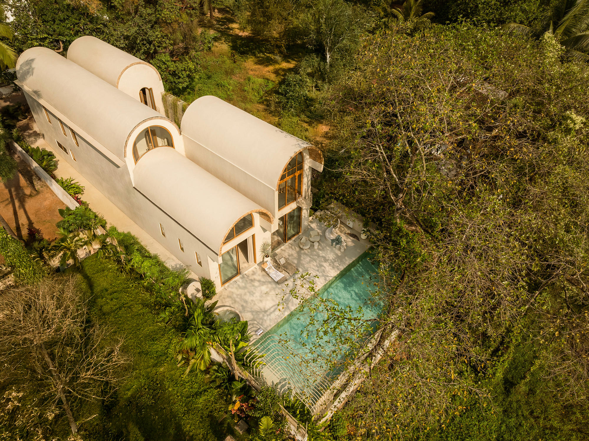 Aerial view of three barrel-vaulted volumes with arched openings beside a pool surrounded by tropical vegetation