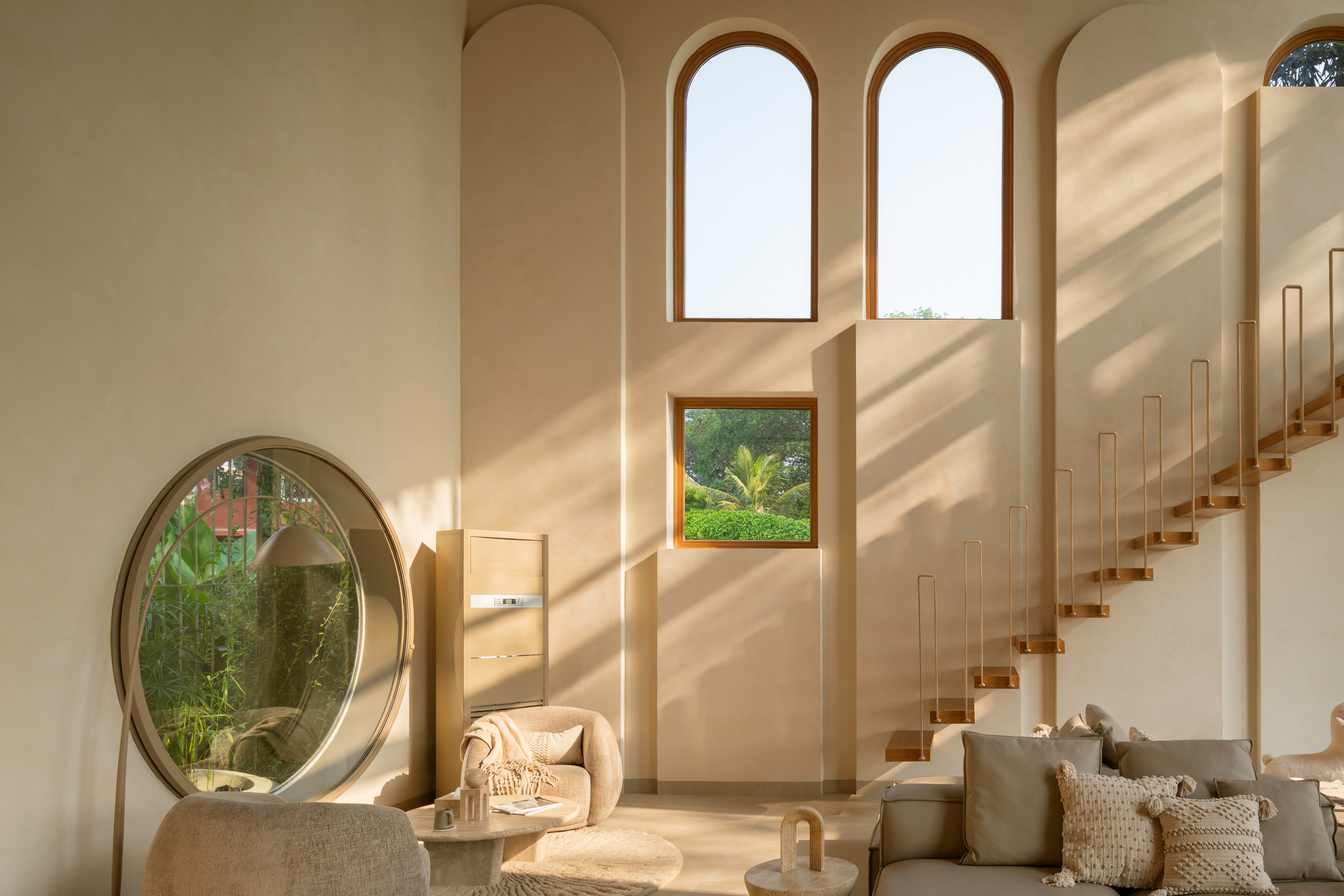 Double-height living room with arched windows, circular portal window, and floating timber staircase in warm sunlight
