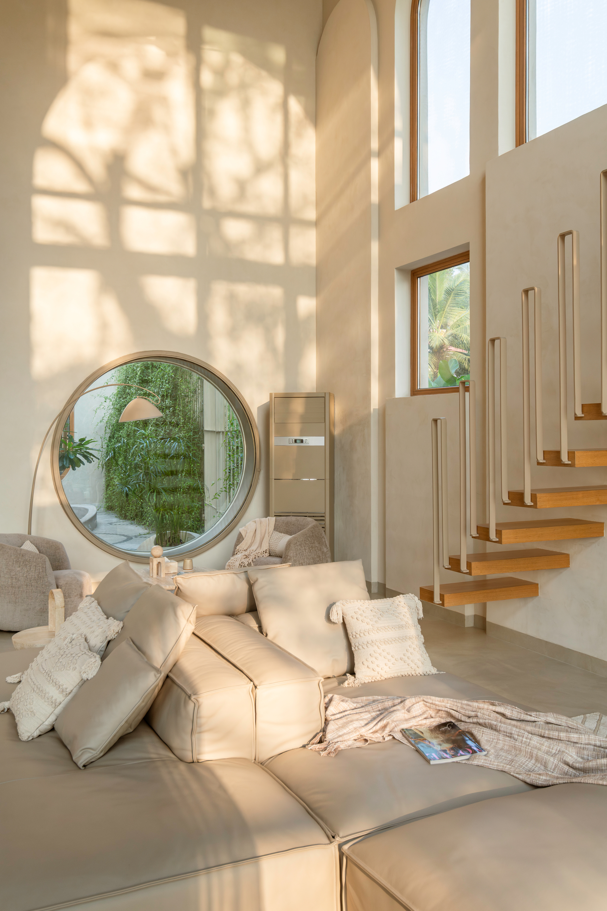 Seating area with circular window looking onto greenery, next to open-riser staircase casting geometric shadows
