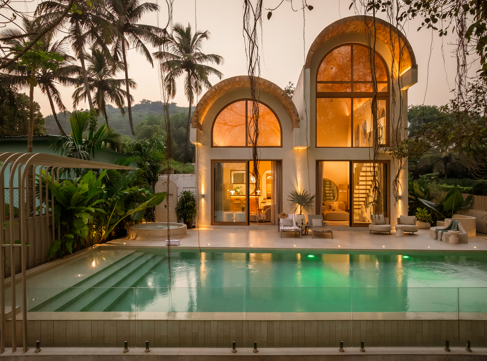 Twin arched glass facades with warm interior lighting overlooking a stepped pool surrounded by palm trees at dusk