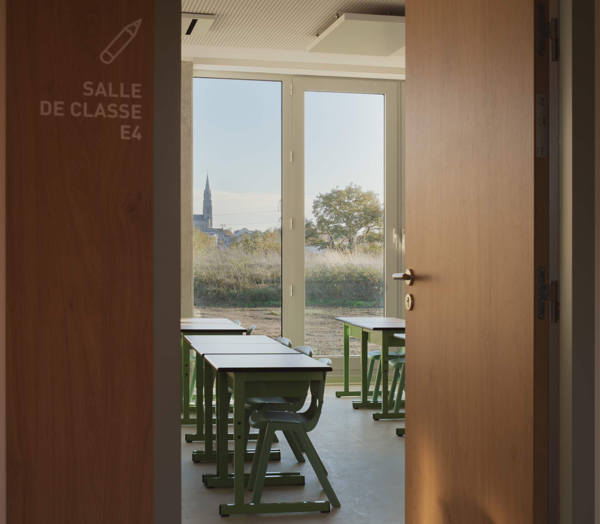 View through open door into classroom with green desks and church spire visible beyond window