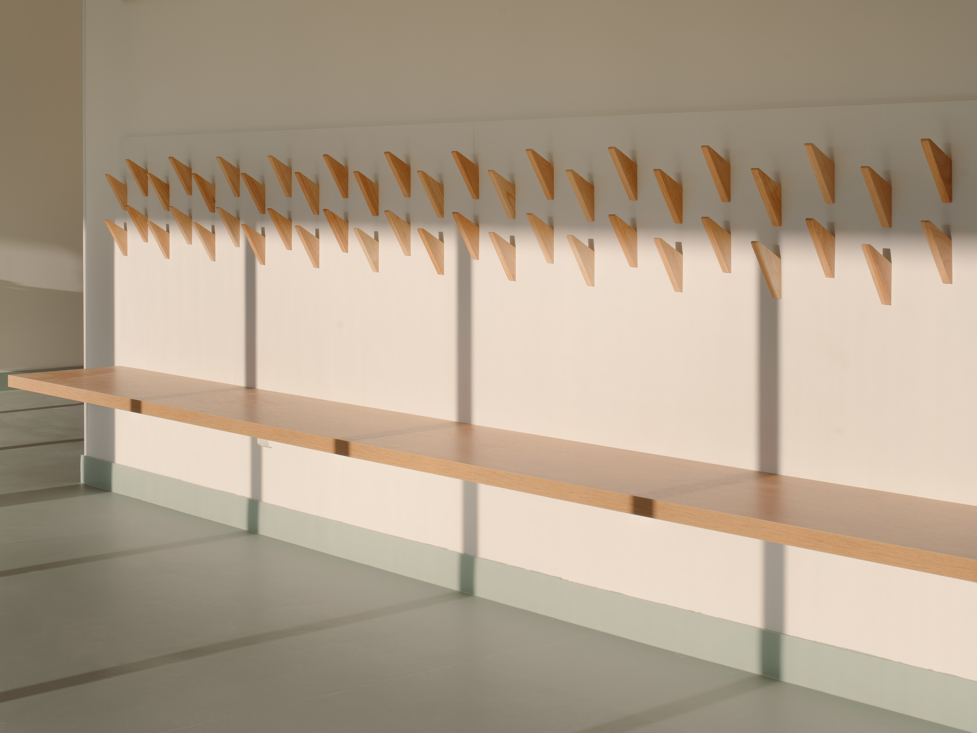 Wall-mounted timber coat hooks arranged in rows above pale green bench in afternoon light