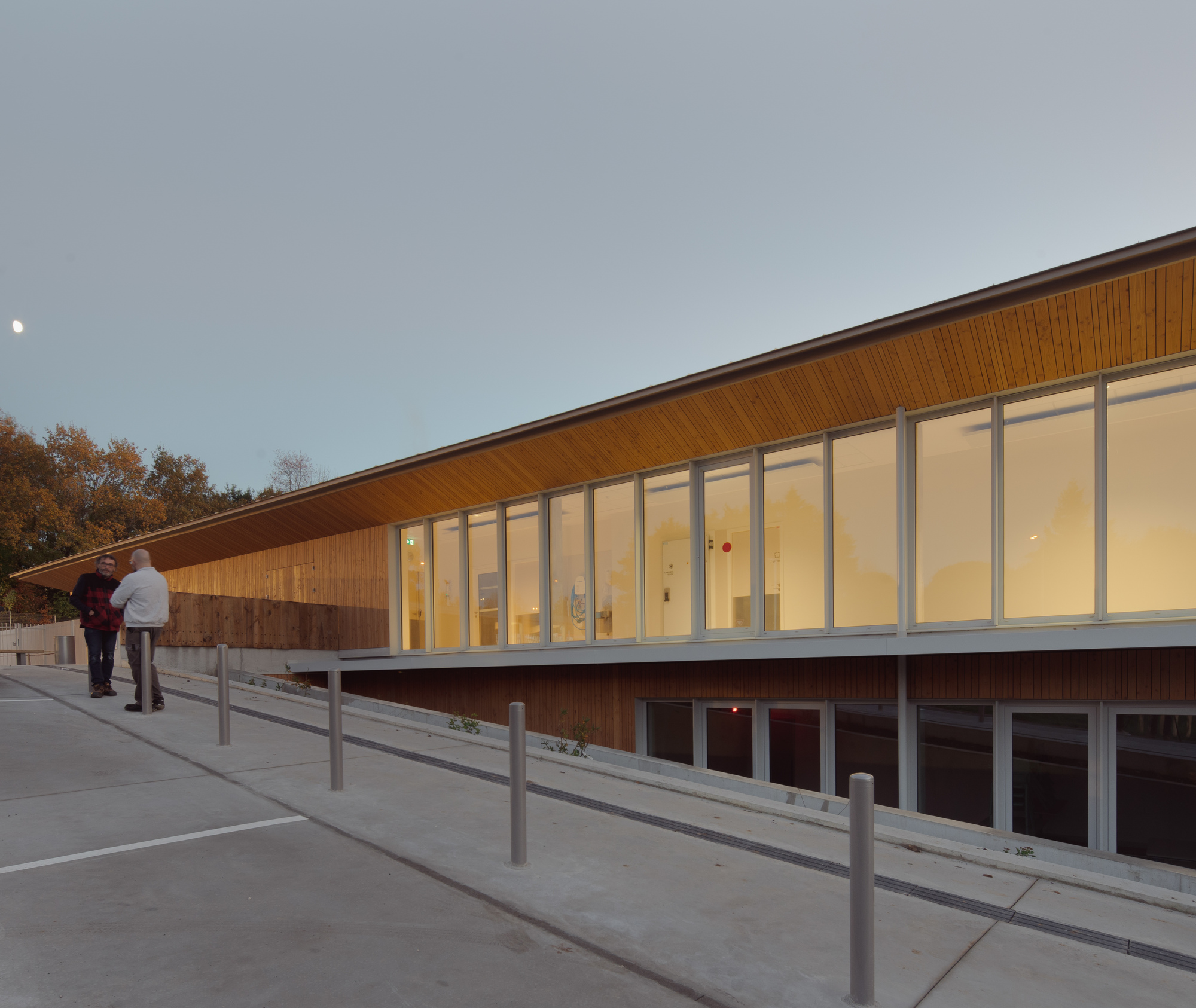 Sloped timber-clad facade with full-height glazing and two figures walking along the exterior ramp at dusk
