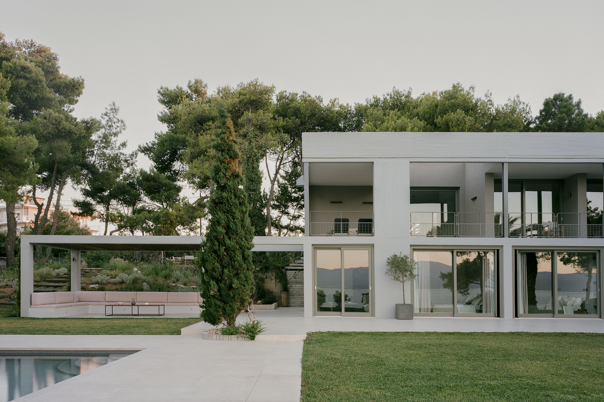 White concrete and glass facade with two-story glazing overlooking a lawn and pool under overcast skies