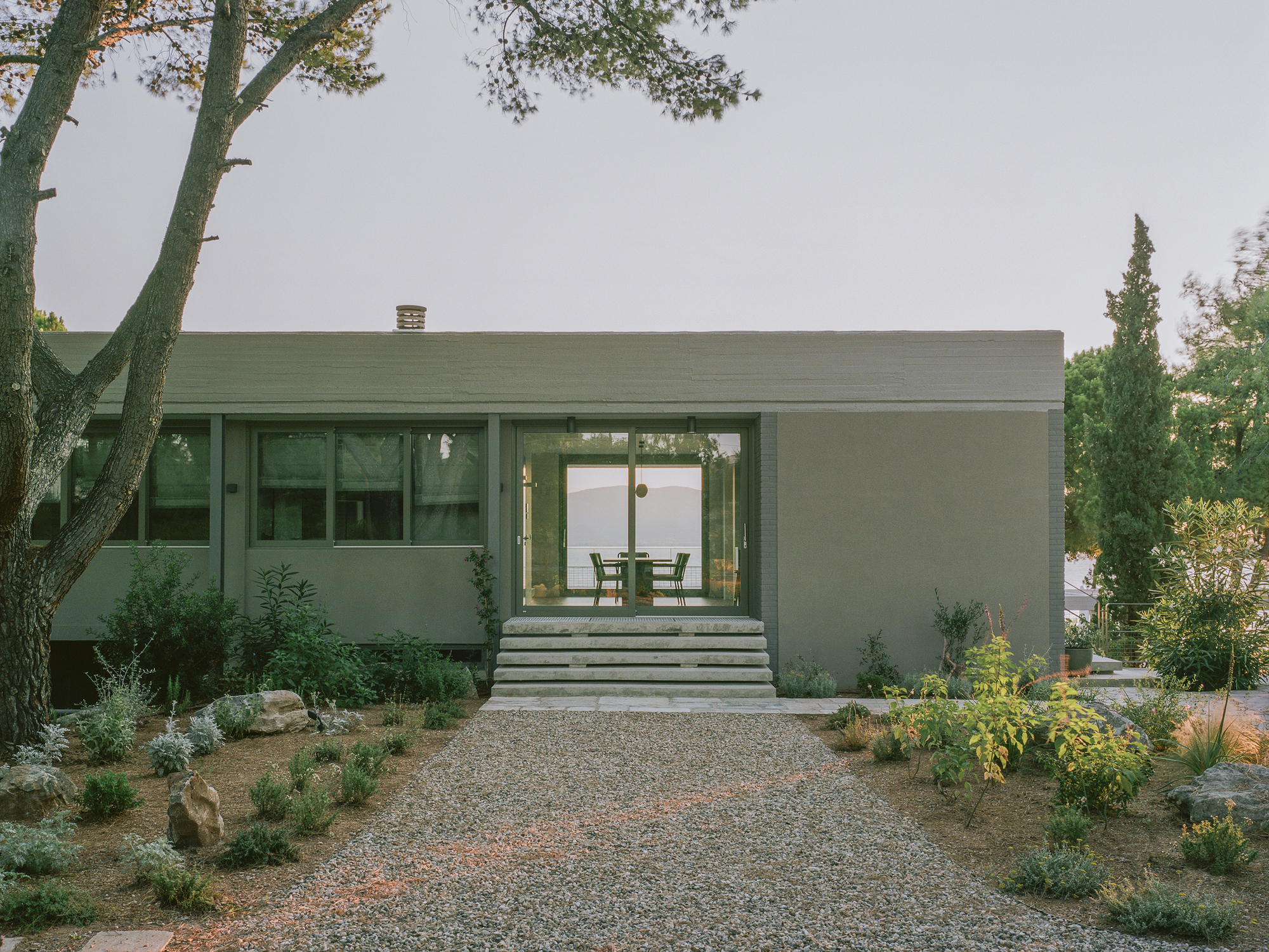 Entry facade with gravel driveway, planted beds, and concrete steps leading through glazed sliding doors