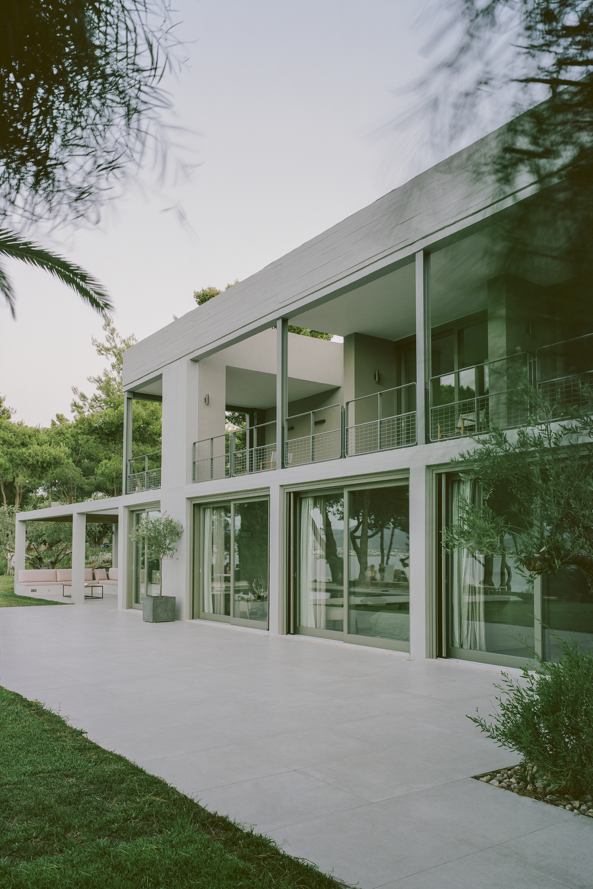 Two-story glass facade with white structural columns framed by lawn and palm fronds overhead