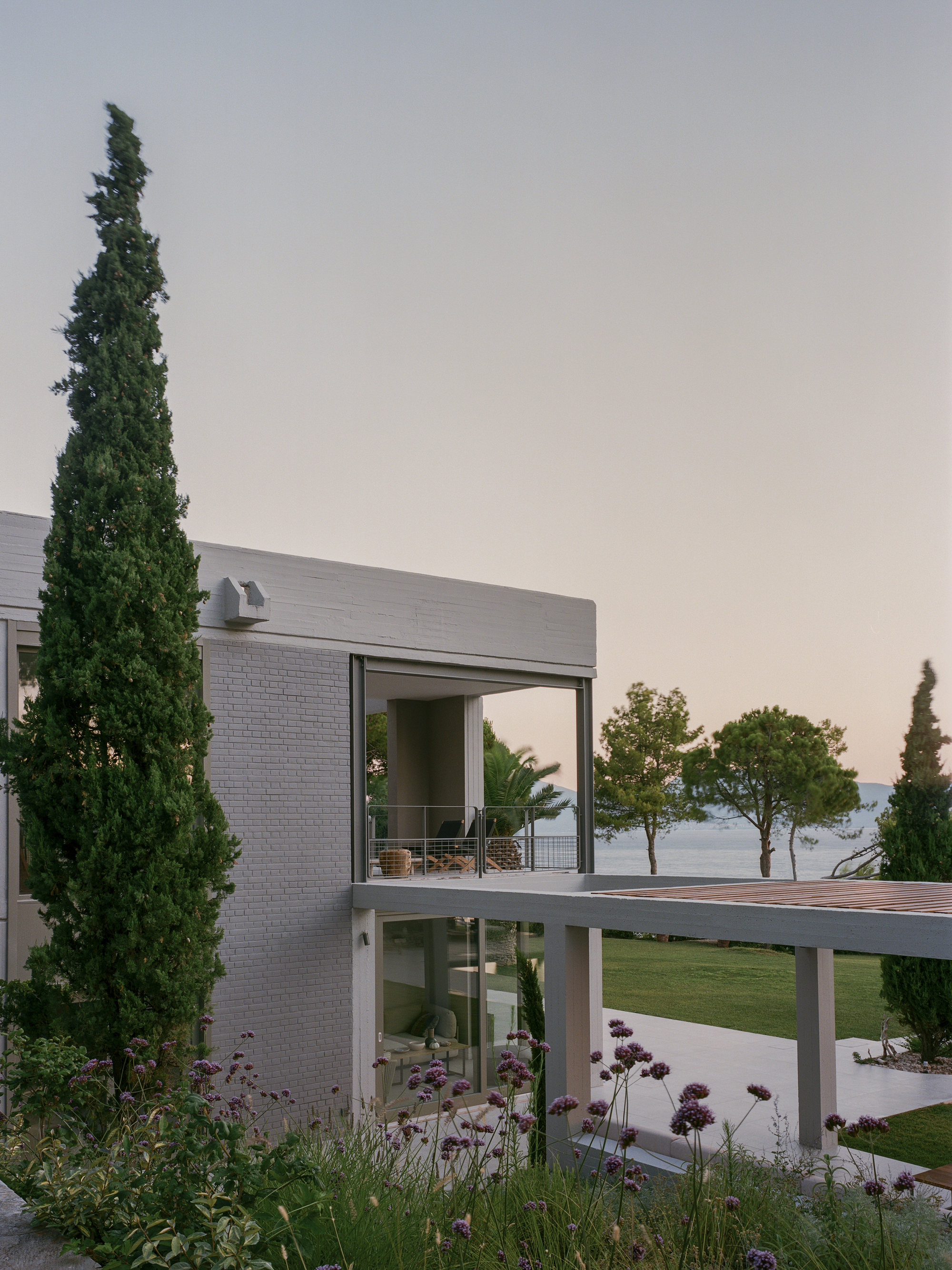 Corner elevation showing grey brick volumes and cantilevered terrace with cypress trees and purple flowering plants
