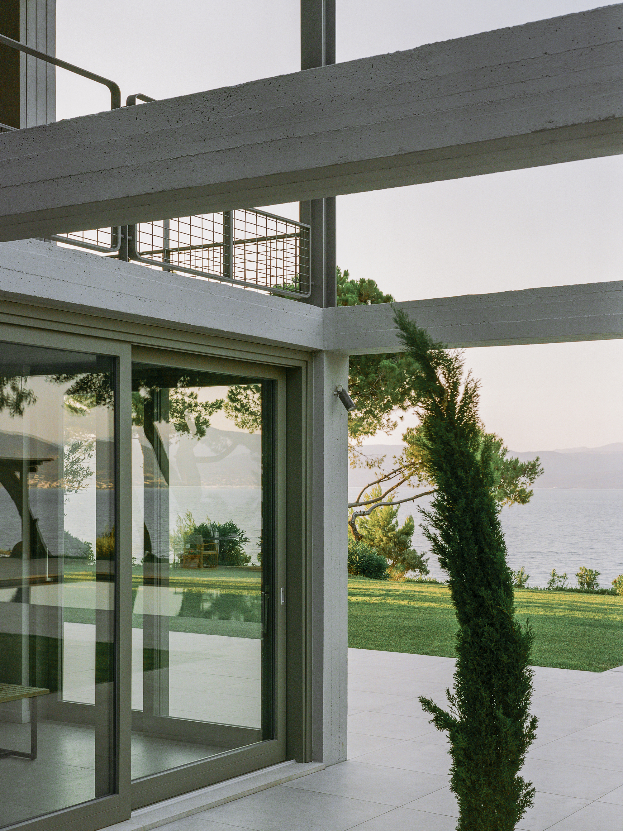 Concrete beams and glazed sliding doors opening to lawn with waterfront and cypress beyond