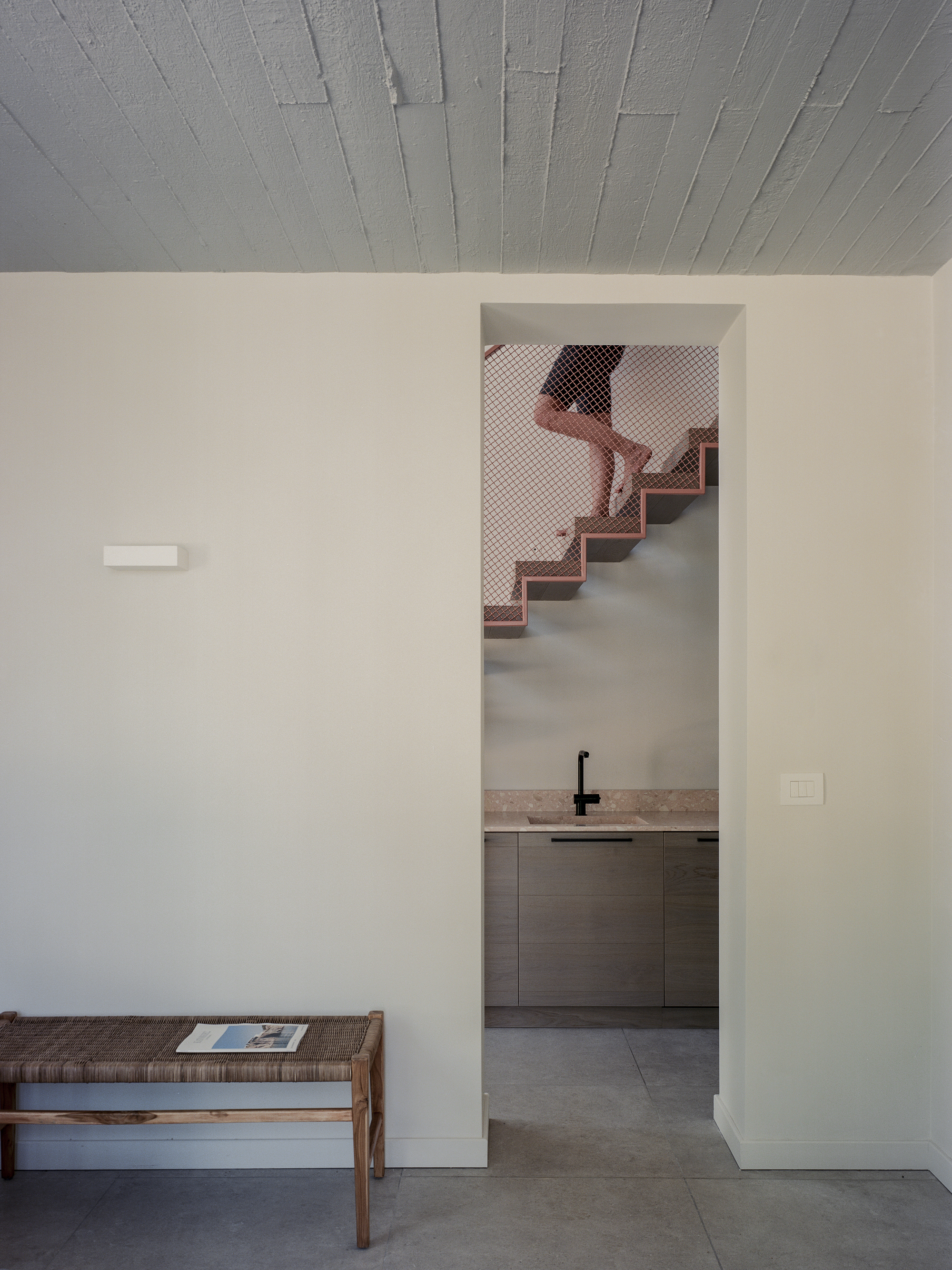 Doorway through a white wall revealing a copper mesh staircase rising beyond a kitchen counter
