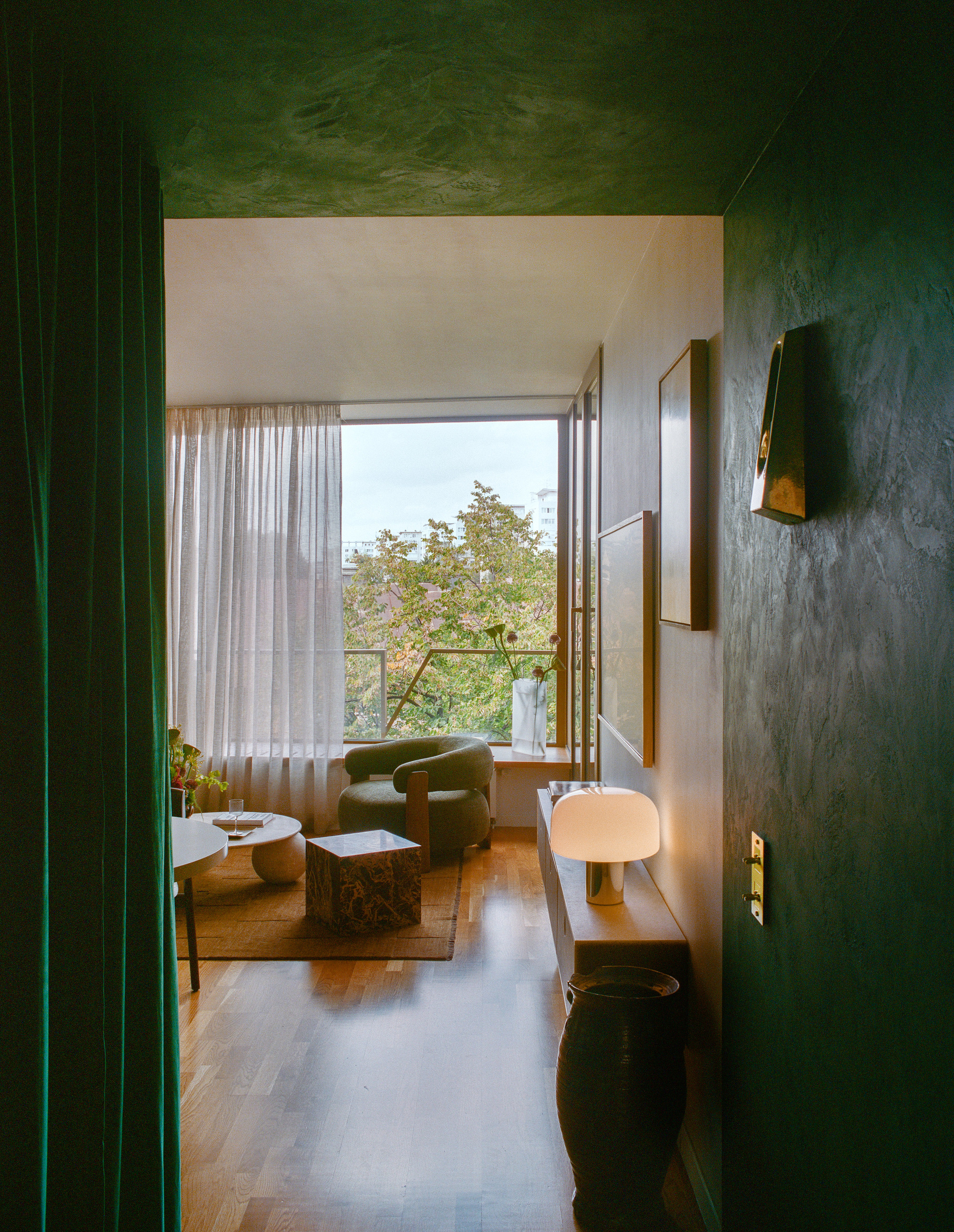 View through green plastered doorway toward a living room with tall windows overlooking tree canopy in daylight
