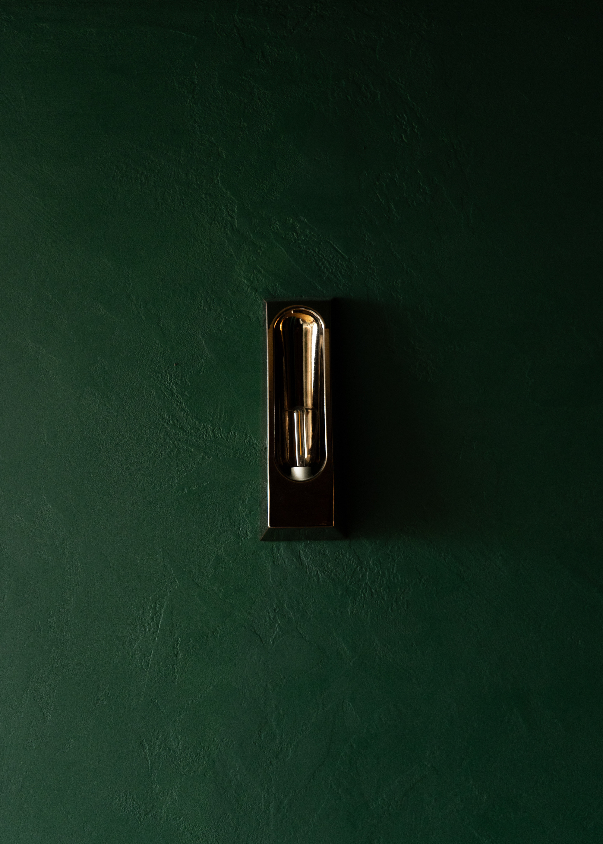 Black wall sconce with exposed filament bulb centered on a deep green plaster wall