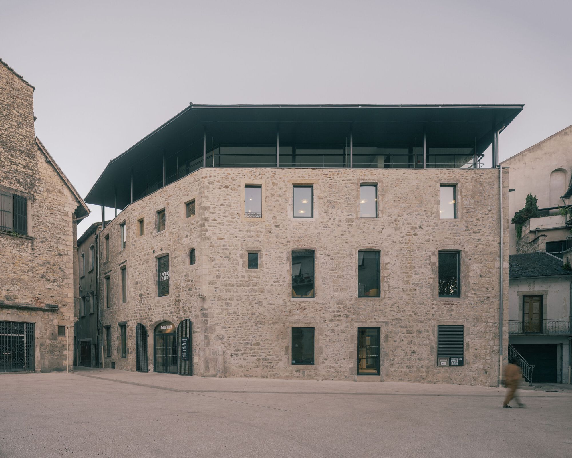 Stone facade with irregularly placed windows and a floating dark roof canopy under overcast skies