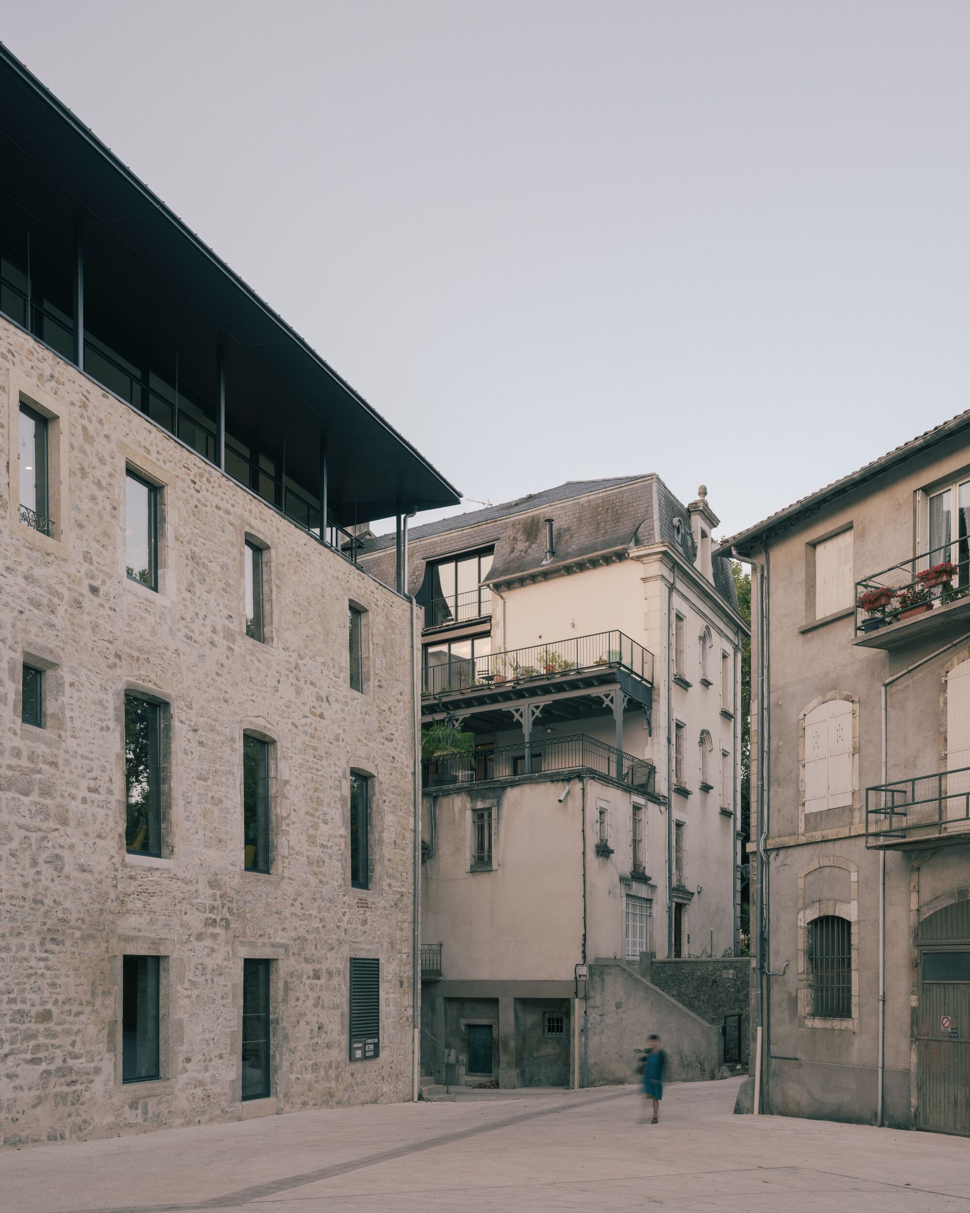 Courtyard view showing restored stone facade with a glazed upper volume topped by a dark metal canopy