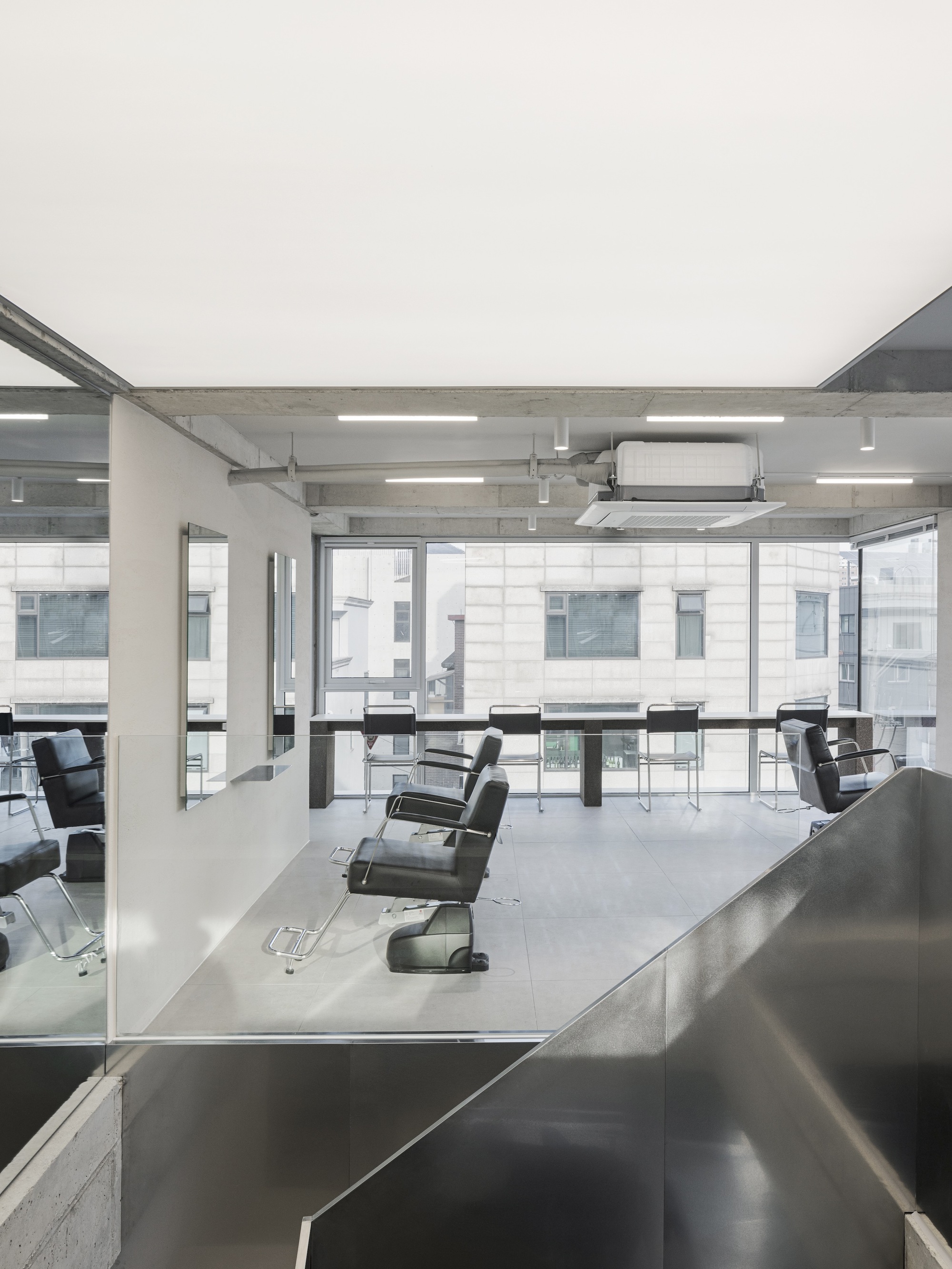 Salon floor with rows of barber chairs facing full-height glazing and exposed concrete ceiling
