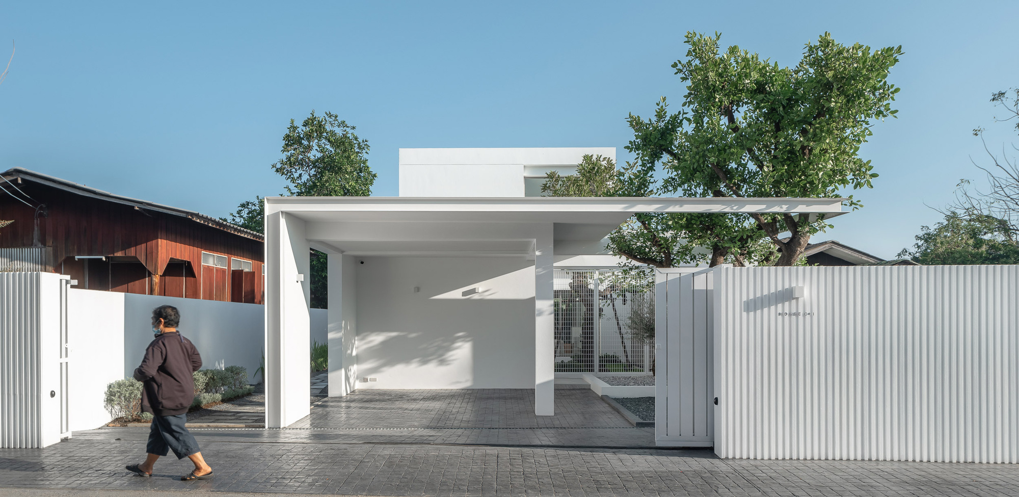 Street facade with white corrugated metal fencing and open carport framing a person walking past