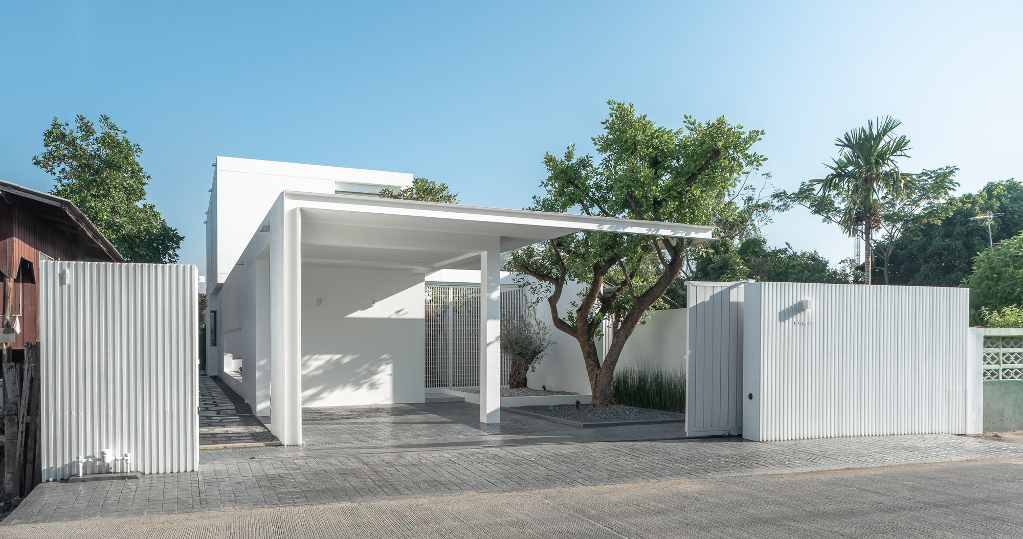 Open carport with tree preserved in courtyard behind white perforated metal wall and paved entry