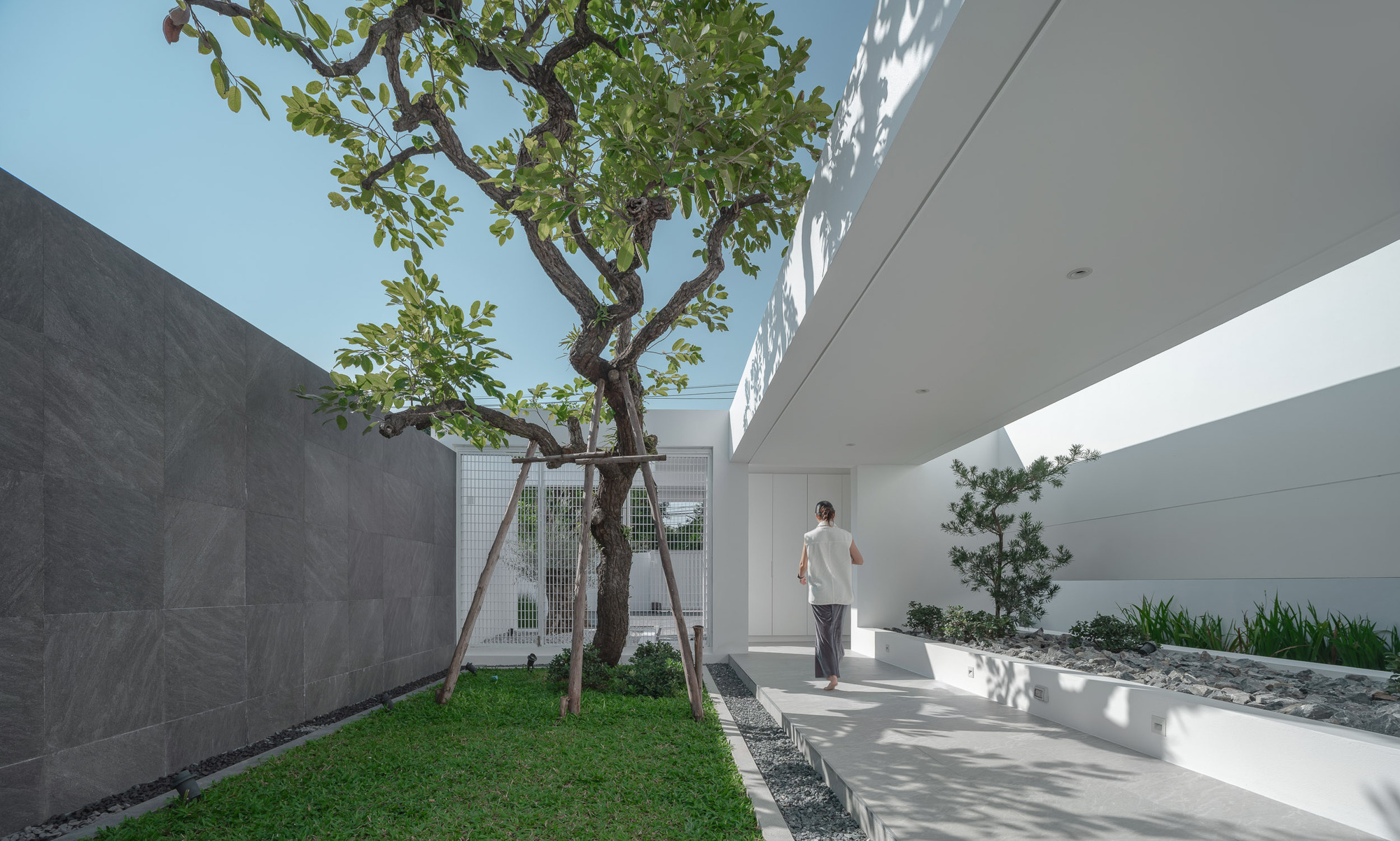 Covered walkway along planted courtyard with concrete walls, glass block screen, and person walking in shade