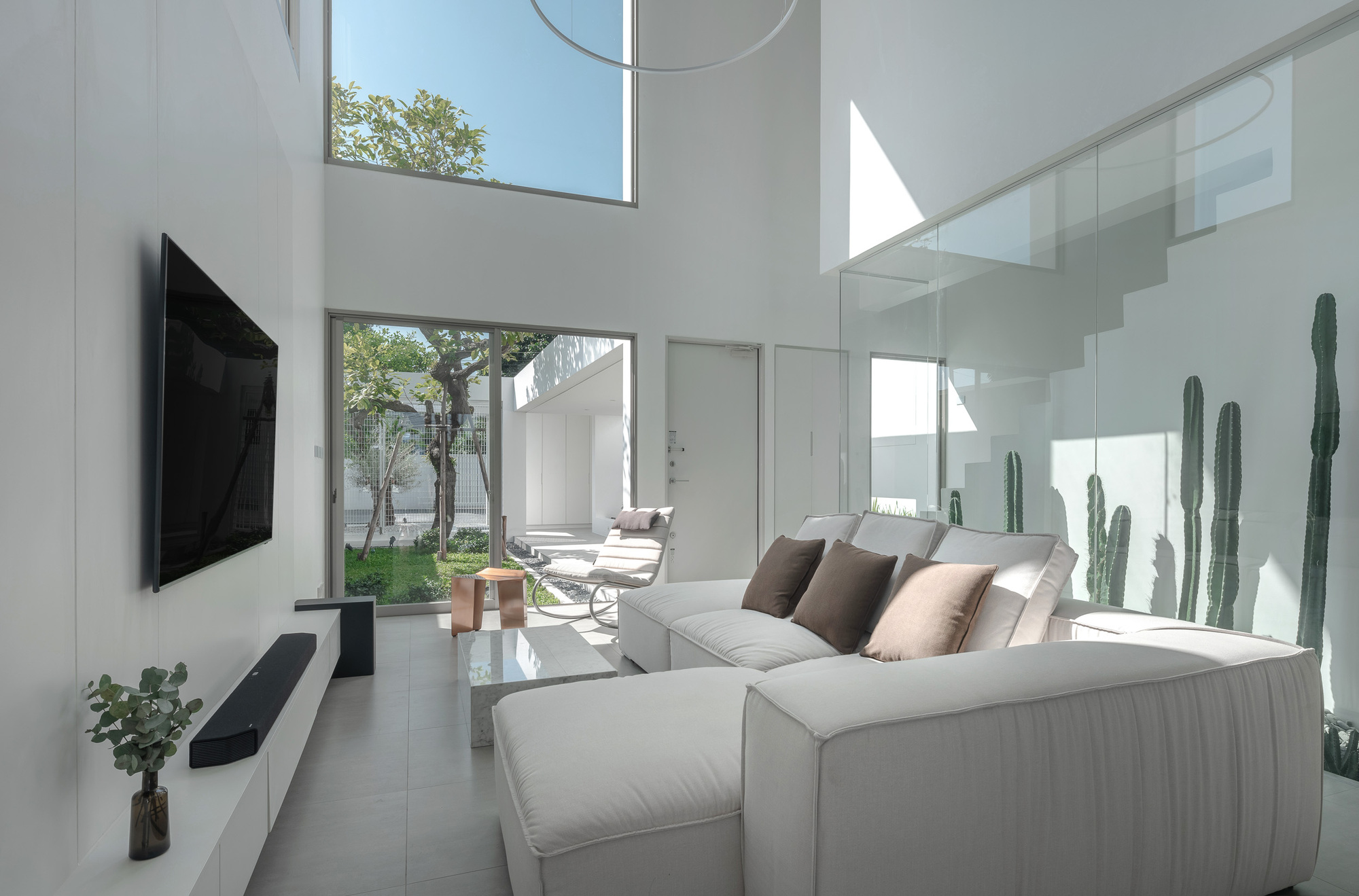 Double-height living room with glass balustrade stair, tall cacti in corner, and view to courtyard beyond