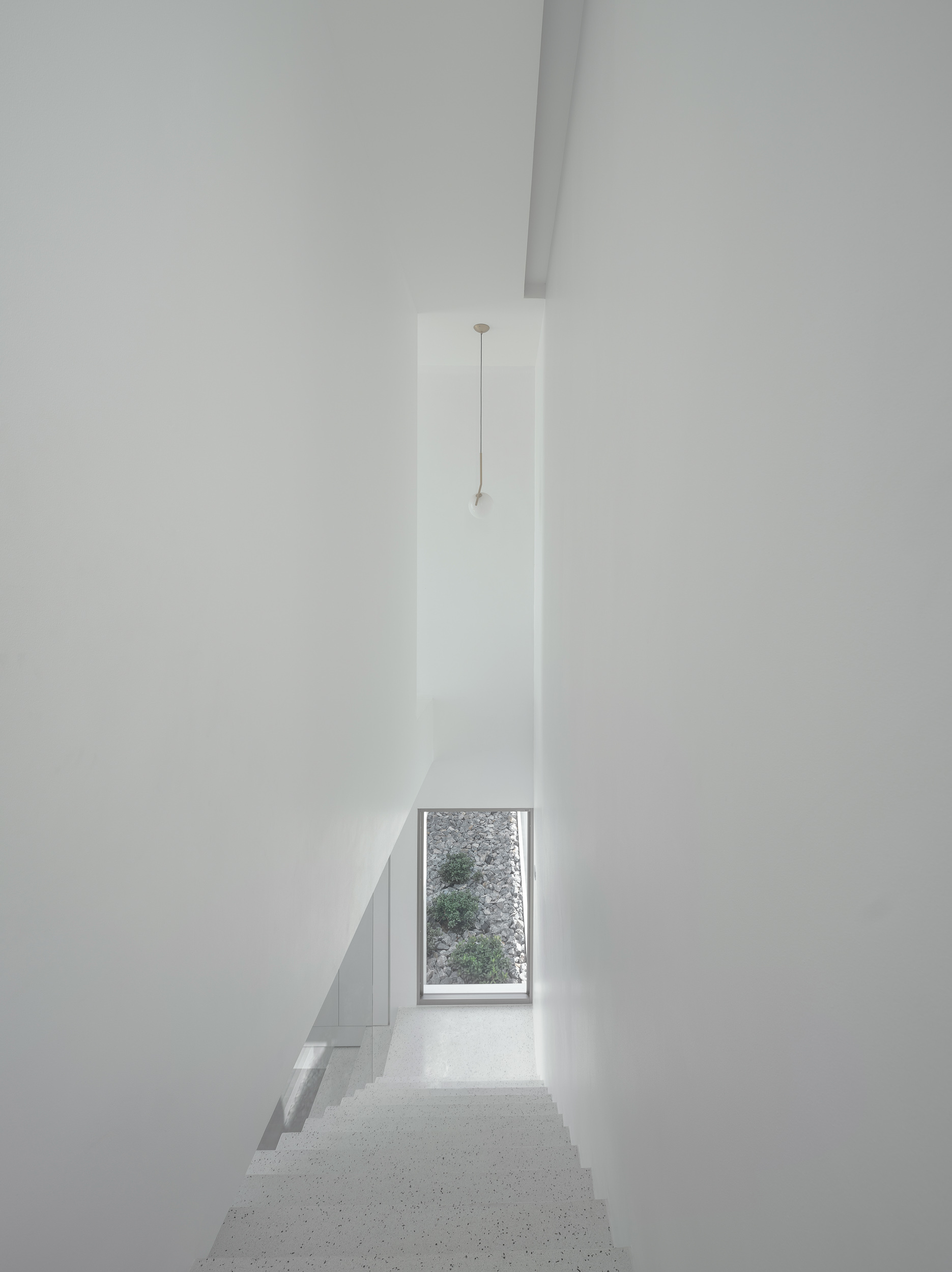 Narrow white stairwell with terrazzo treads descending toward a tall window framing greenery