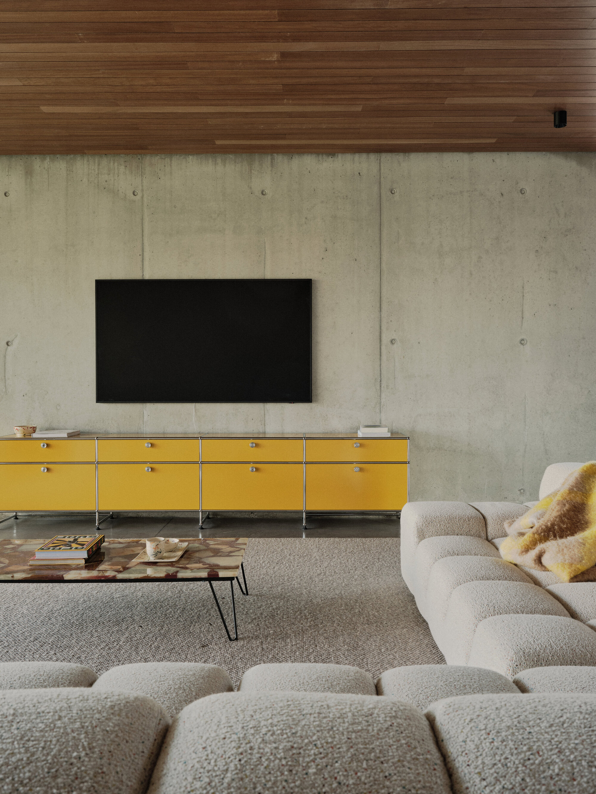 Living room with board-formed concrete wall, timber ceiling and yellow modular credenza beneath flat screen