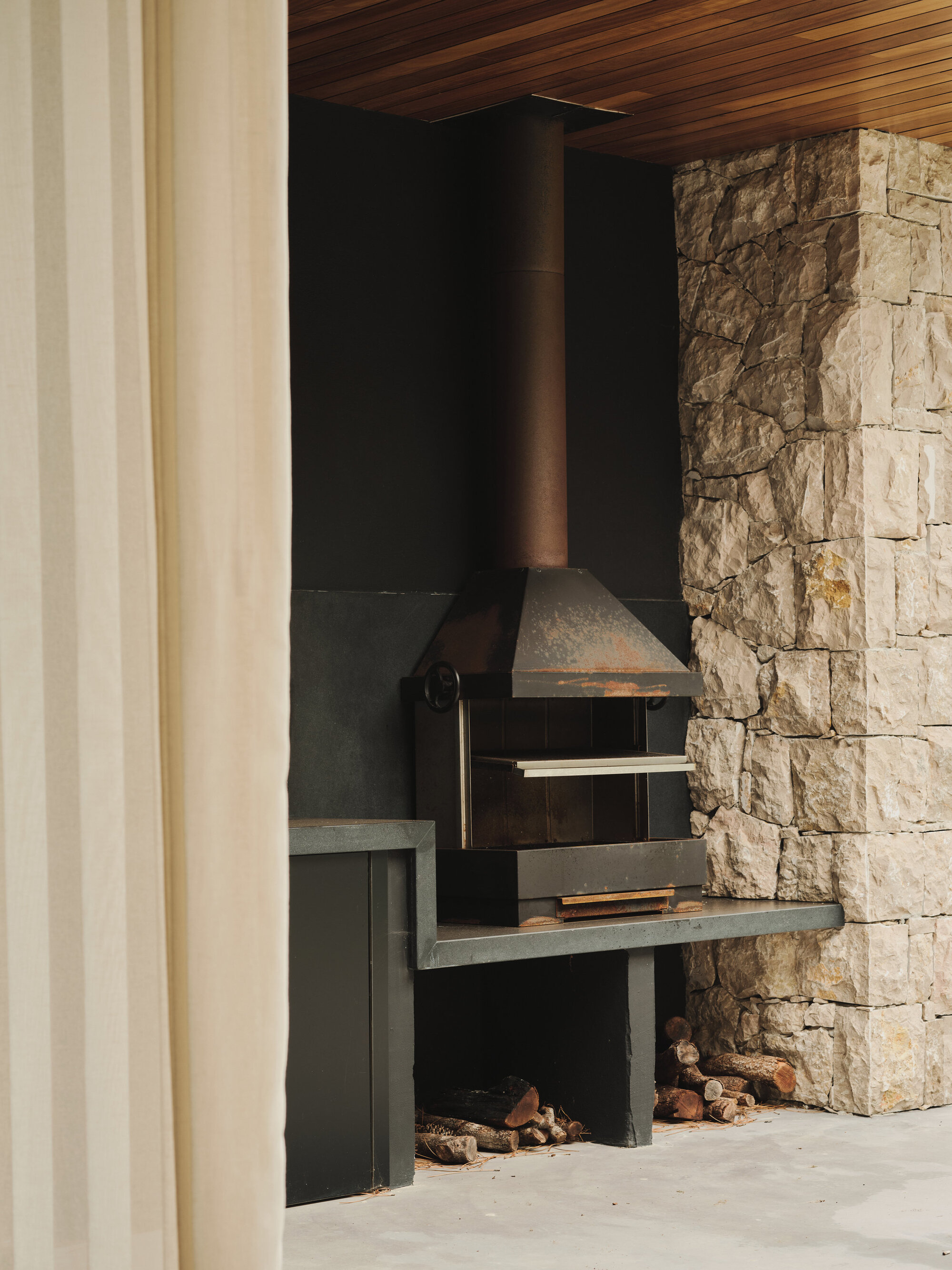 Covered outdoor barbecue area with stone wall, timber ceiling and metal grill under warm afternoon light