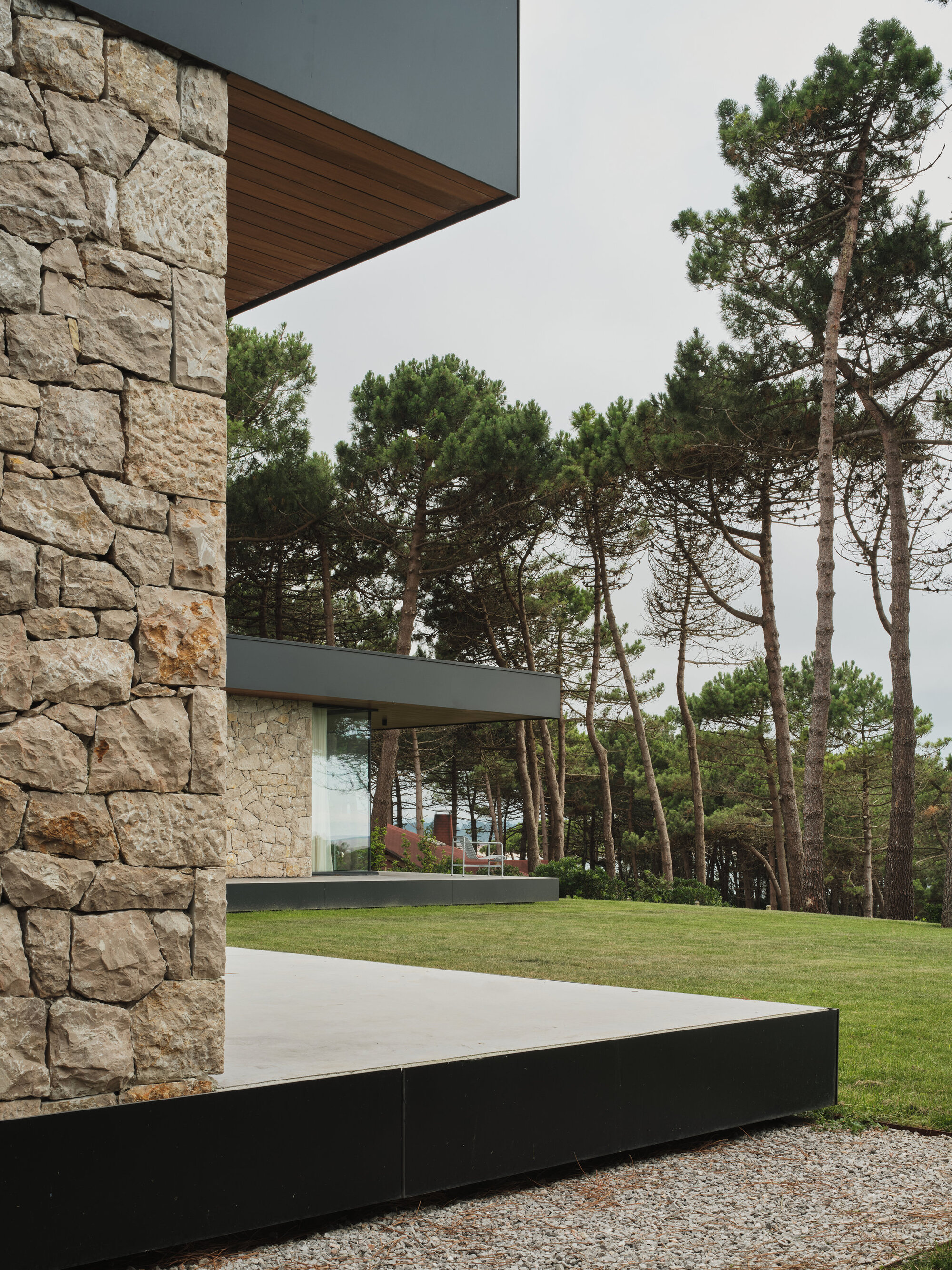 Stone tower with timber soffit and black plinth platform surrounded by pines and gravel