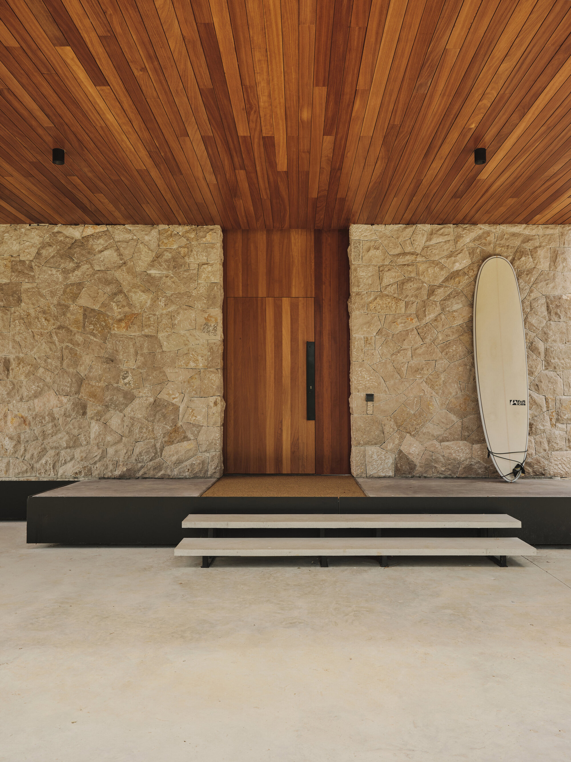 Entry sequence with timber door, striped ceiling, stone walls and surfboard leaning beside wooden bench