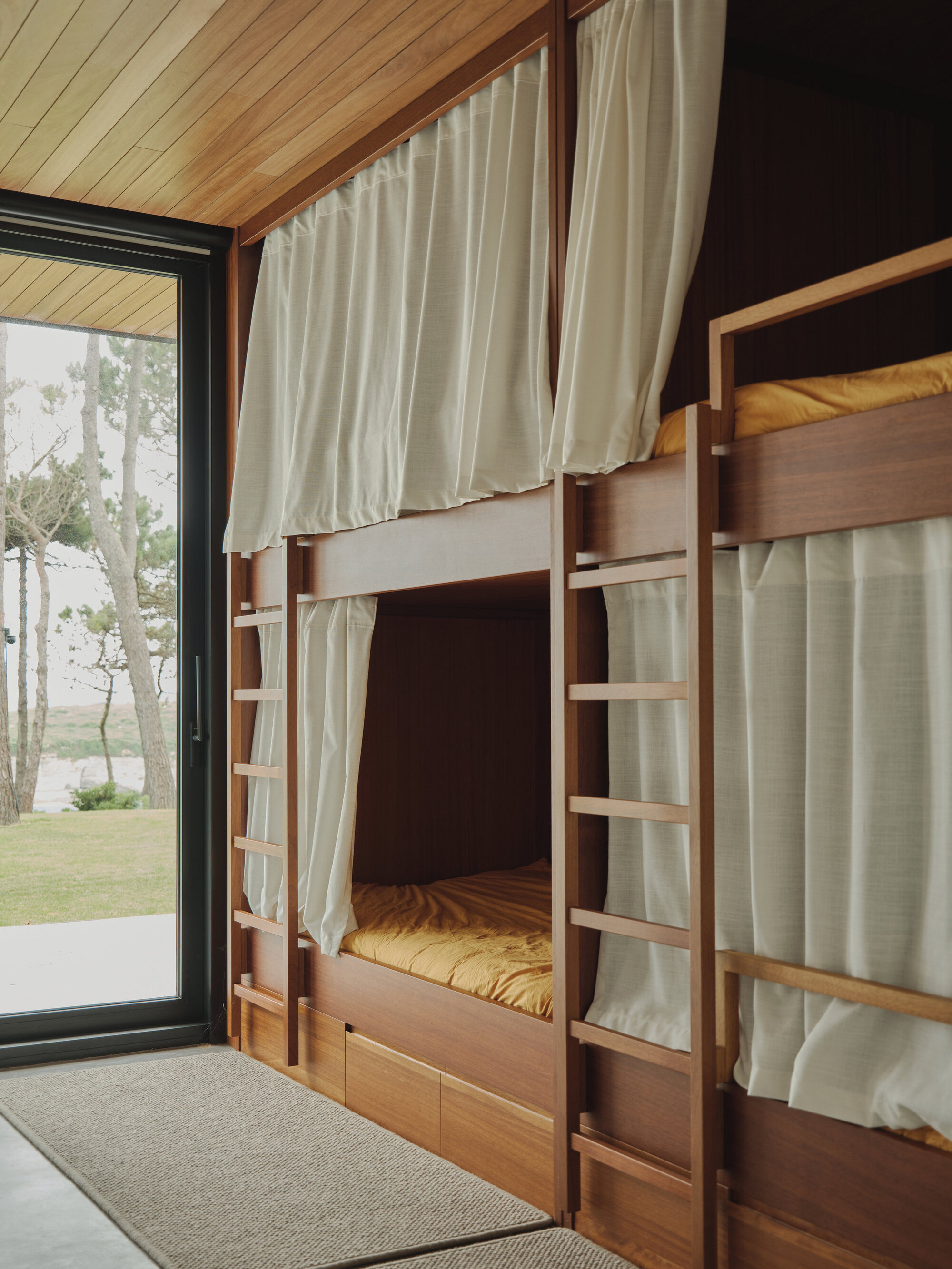 Built-in timber bunk beds with linen curtains and ladder beneath a pitched ceiling with forest views beyond