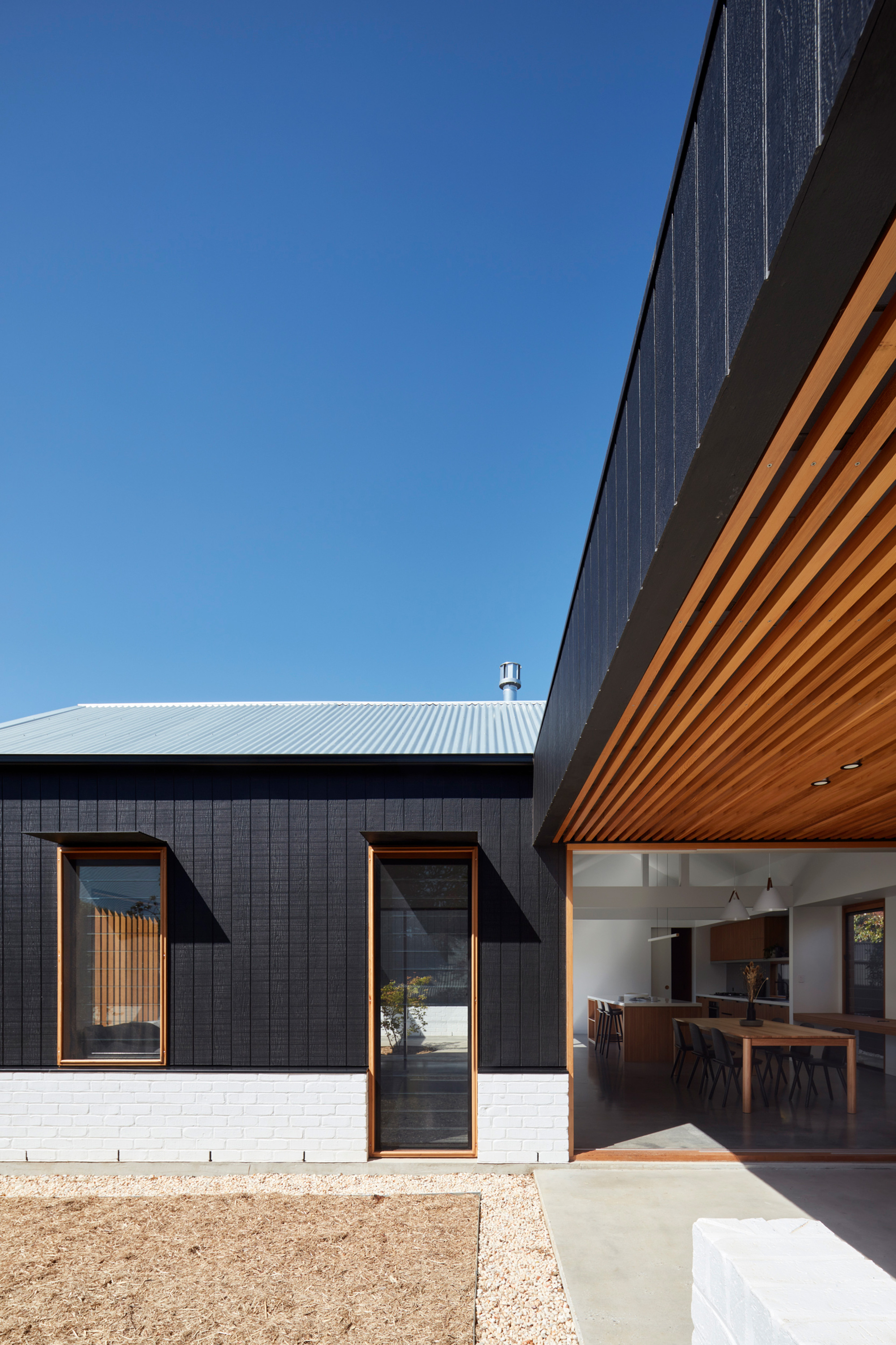Exterior courtyard view showing charcoal cladding, white brick base, and timber soffit under clear blue sky