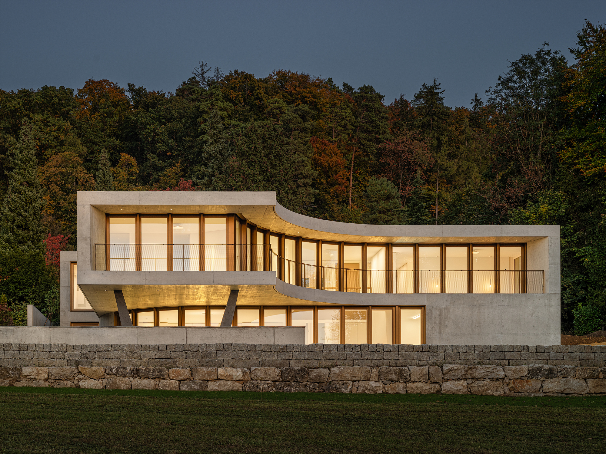 Concrete and timber facade with undulating roofline illuminated at dusk behind stone retaining wall
